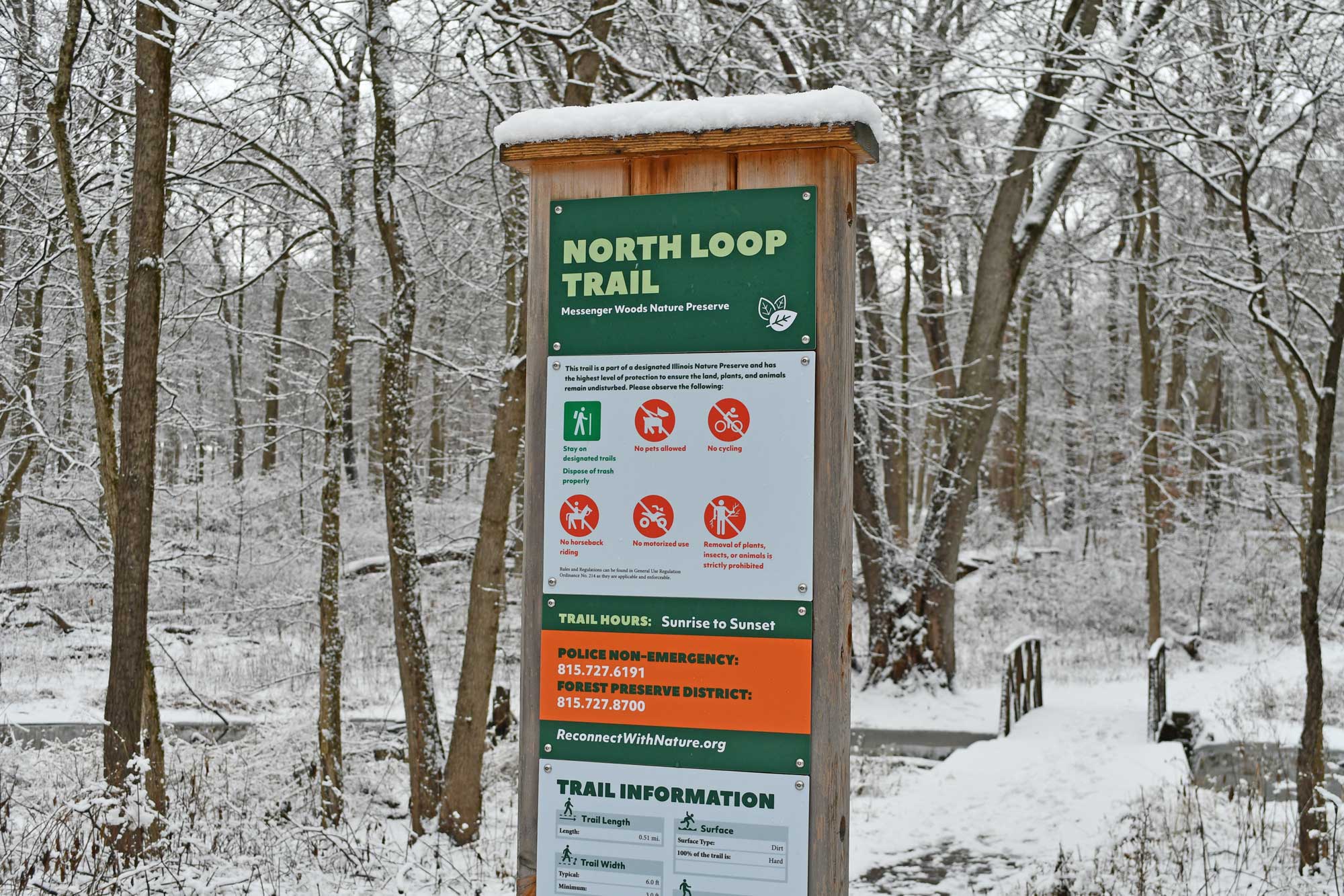 Trail sign with snow covered trail and trees in the background.