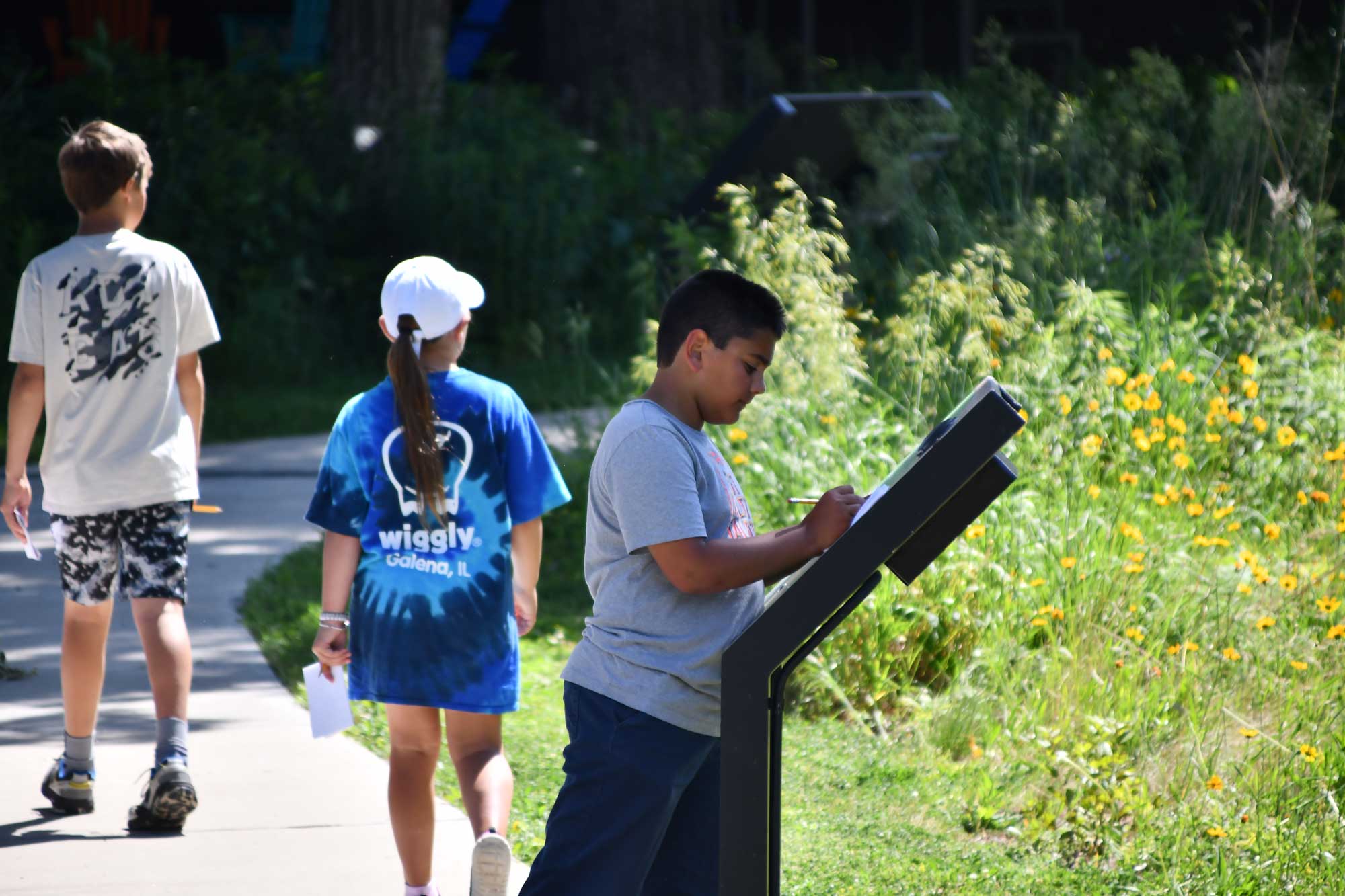 A child standing at an interpretive sign with two kids on a trail in the background.