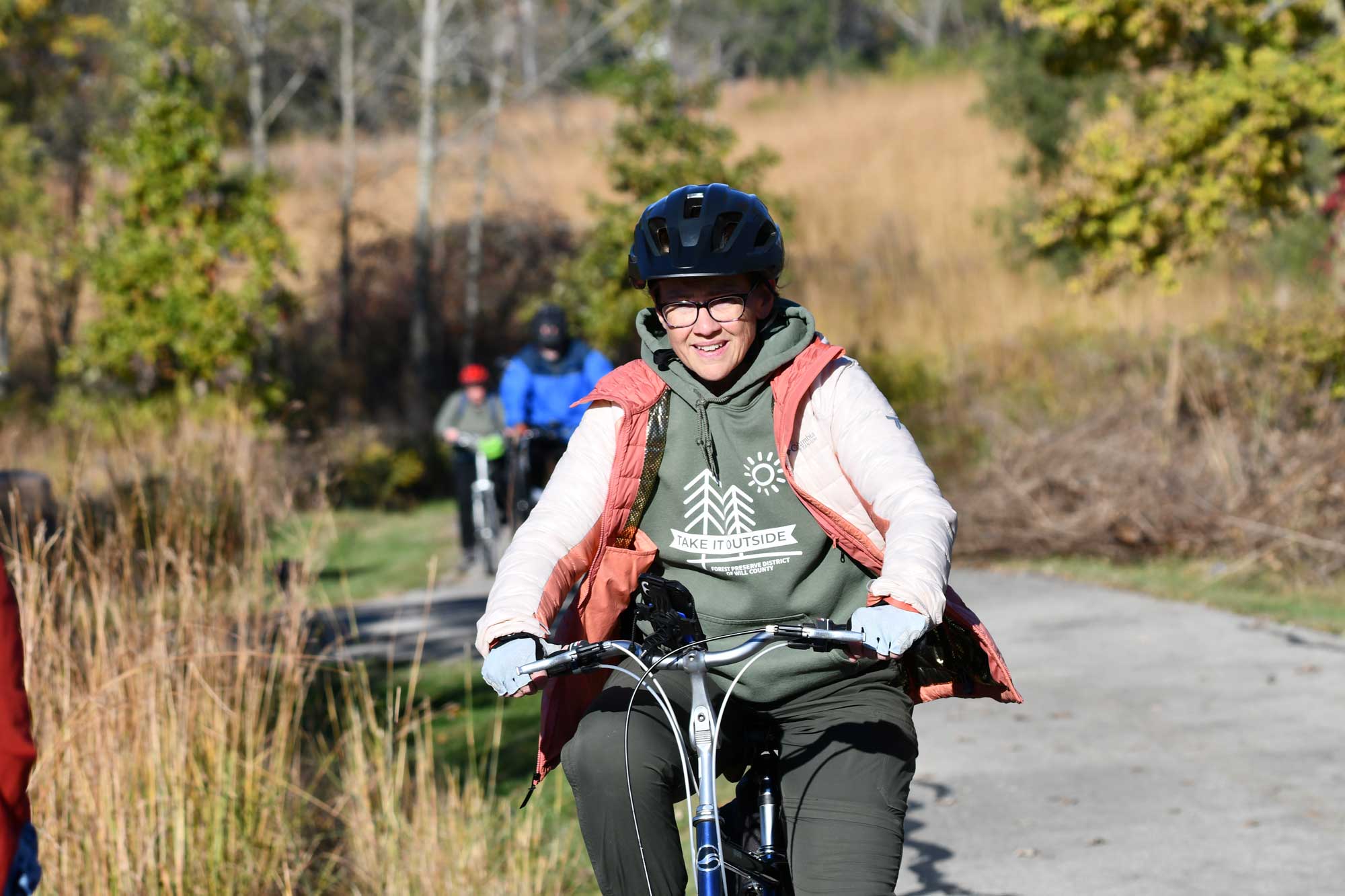 Person riding bike on a paved trail with other bikers and vegetation in the background.