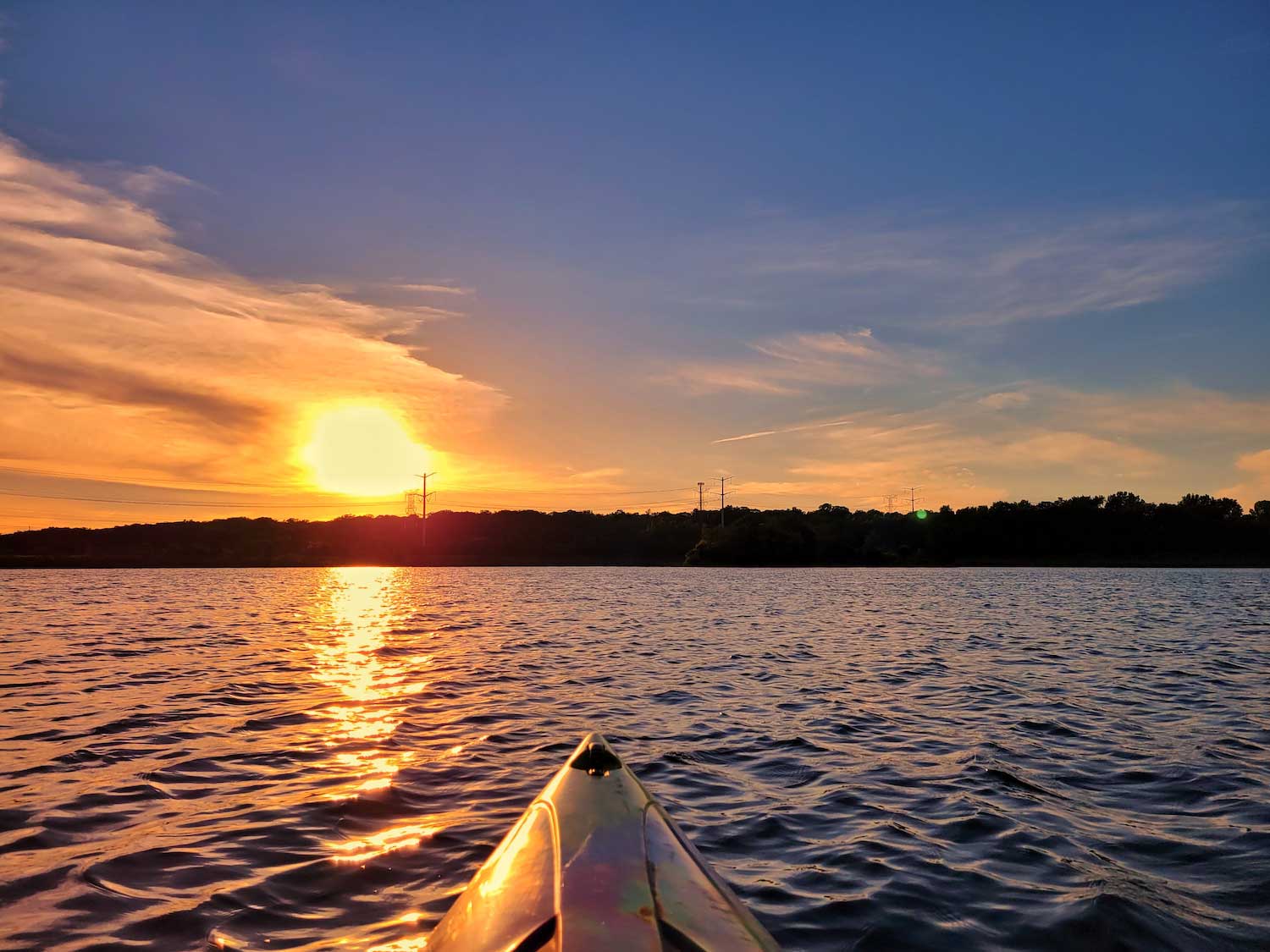 The orange sun in the distance over water with trees lining the shore. The tip of a kayak is visible in the water.