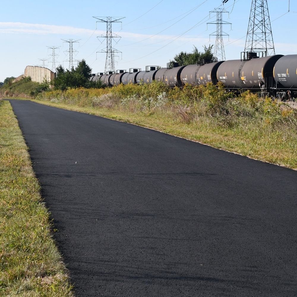 A paved trail runs alongside a railroad track lined with black freight train cars and tall power line towers.