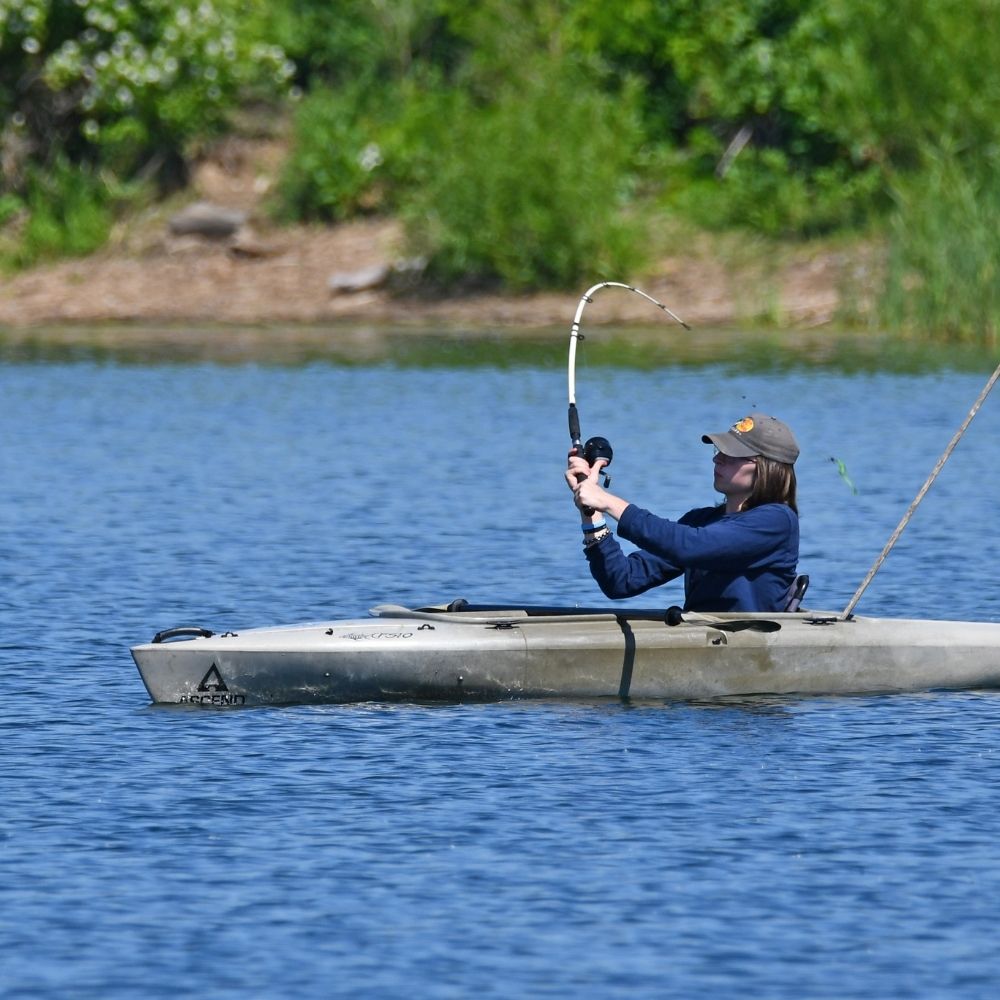 A person in a kayak fishes on a calm lake, holding a bent fishing rod that suggests a catch on the line.