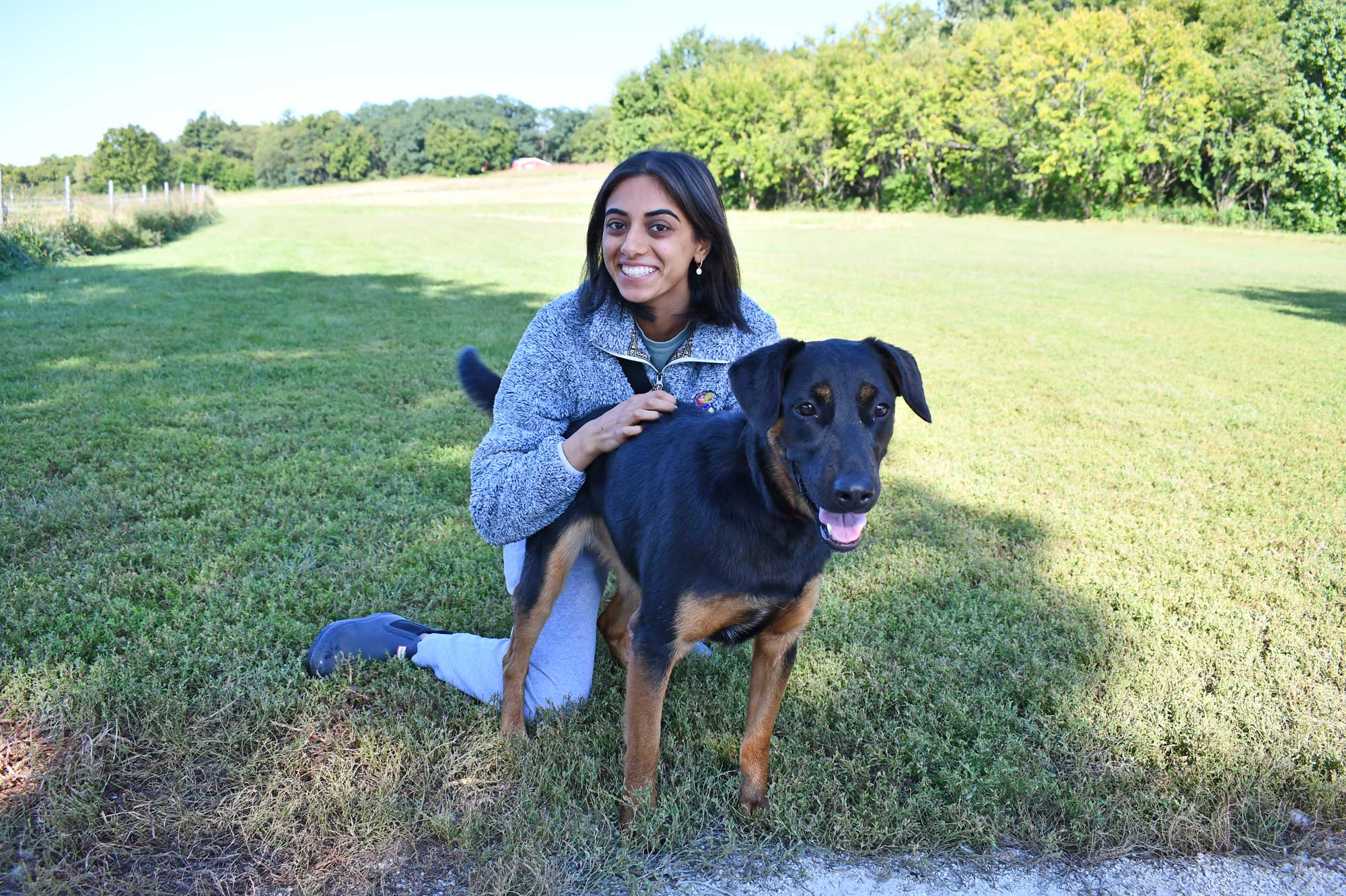A person poses with her black and brown dog in a grassy open area.