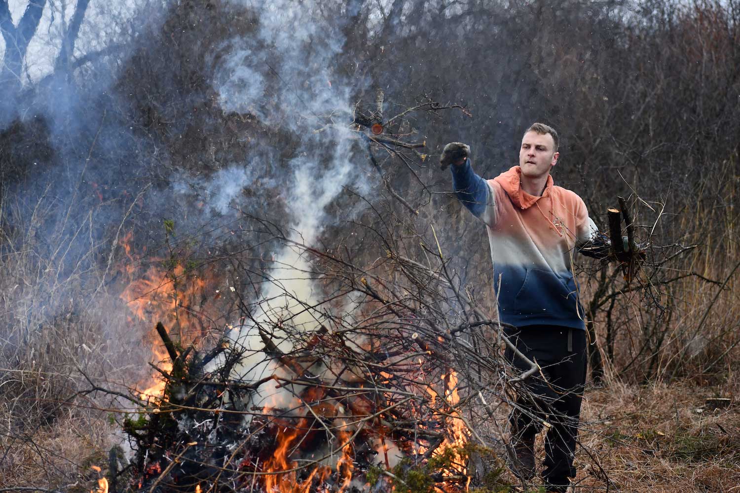 A young man adding branches to a burning brush pile.