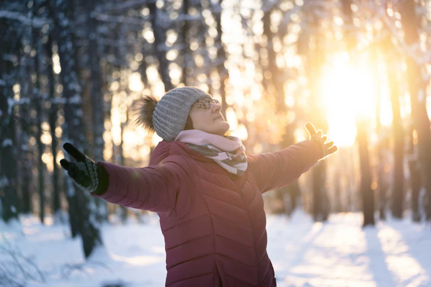 A person standing with arms extended in a snowy forest with the sun peeking through the trees.