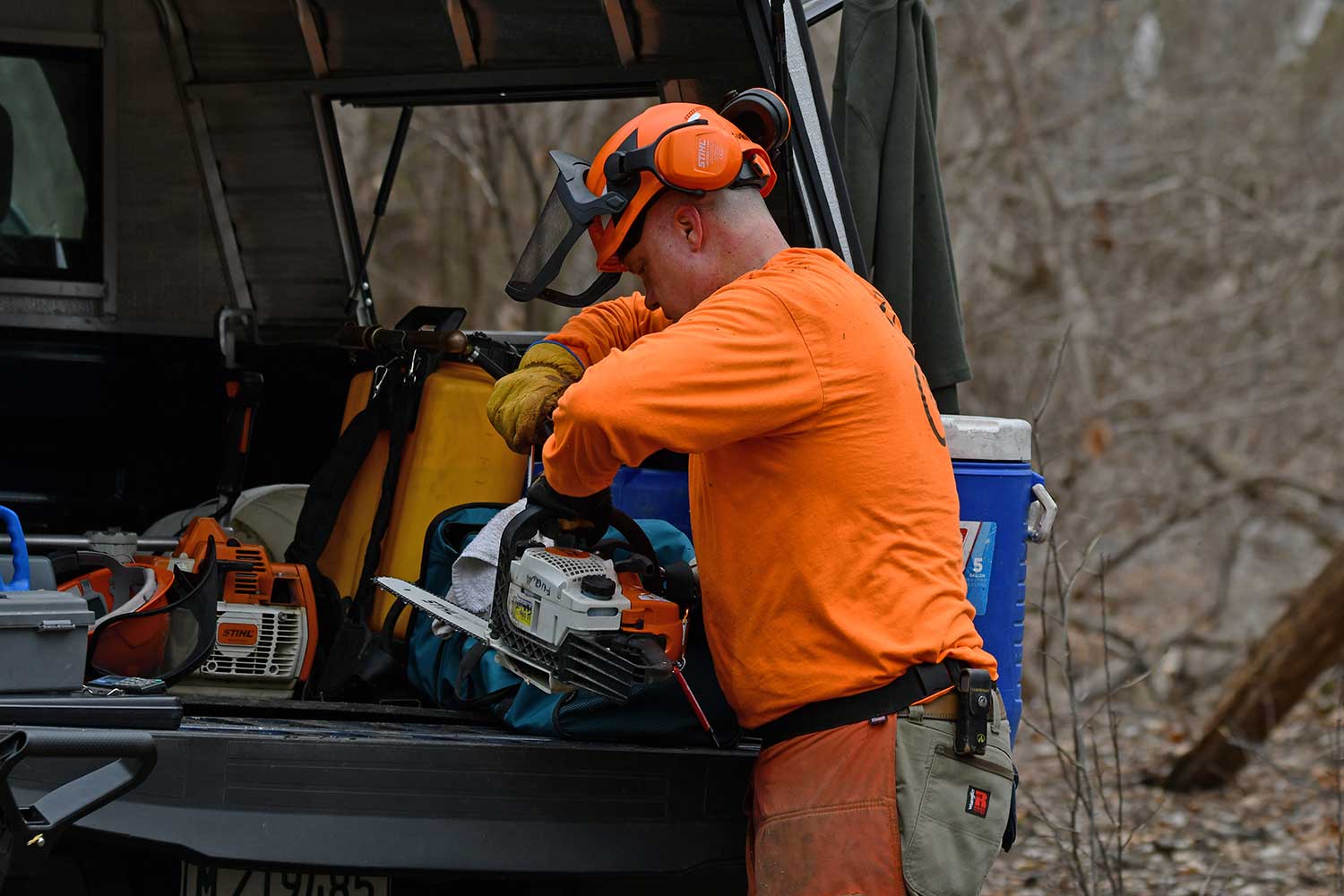 A man working on a chainsaw at the back of a pickup truck loading with gear.