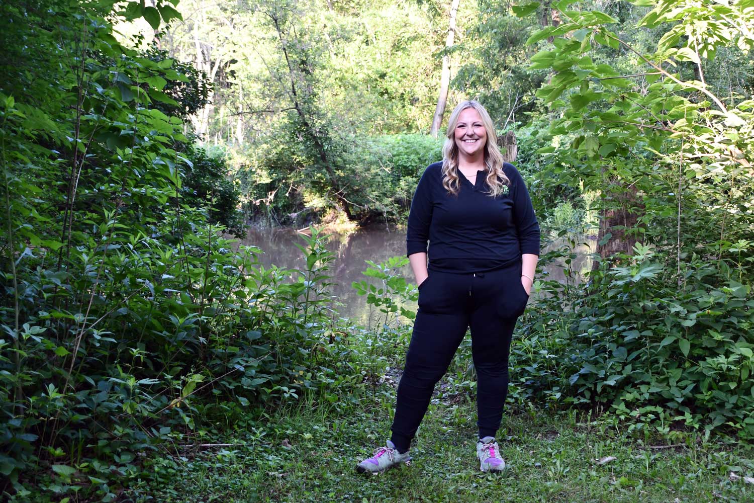 A woman standing on a trail along a wooded creek.