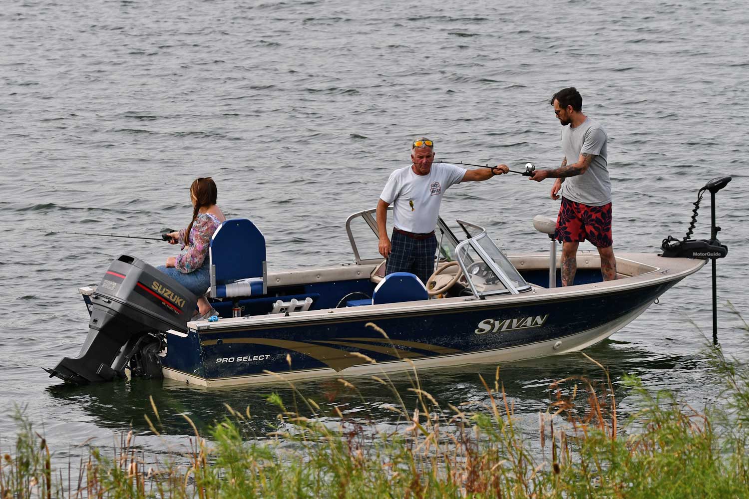 Three people in a boat fishing near some tall grasses.