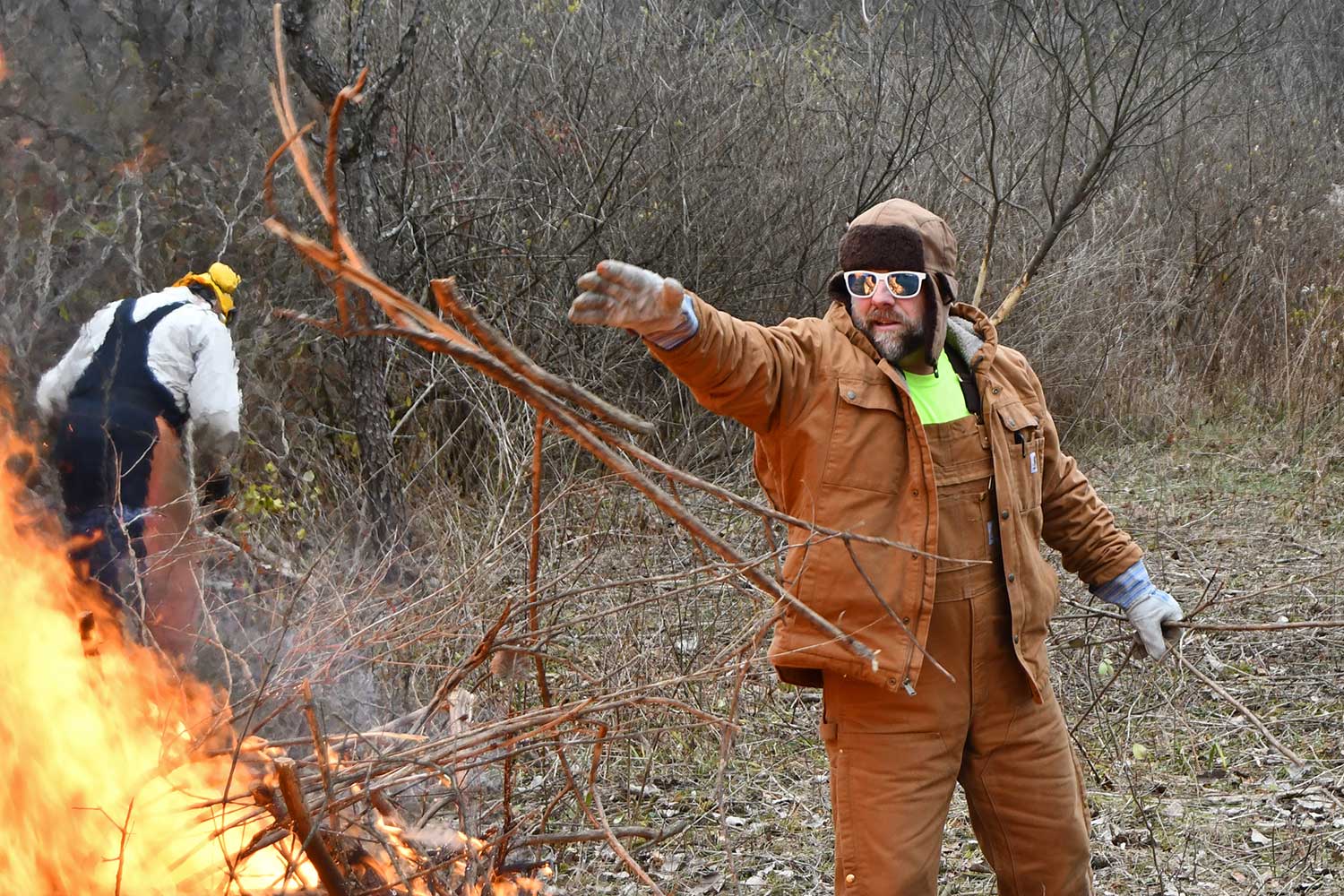 A person throwing brush on a burn pile while a person works in the background.
