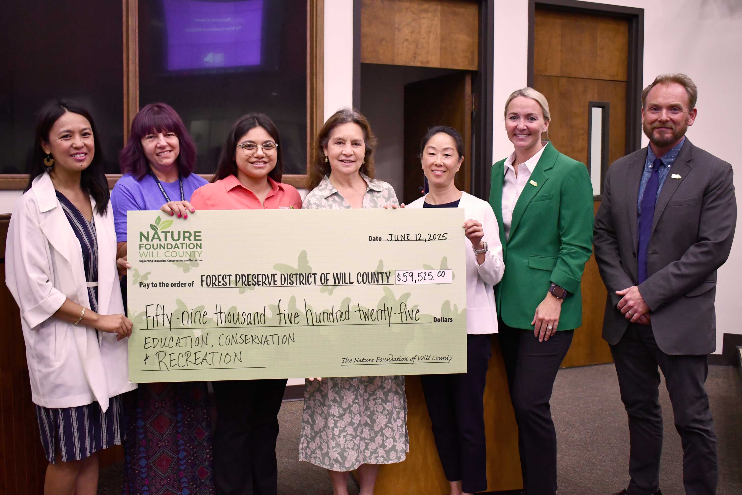 A group of seven people stand together in a conference room, smiling and holding a large ceremonial check. 