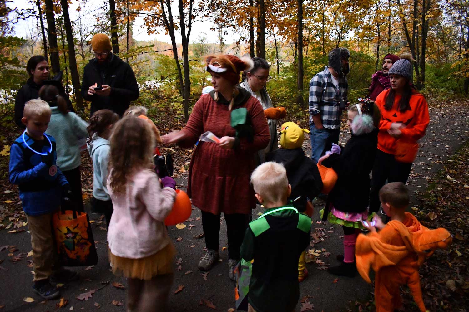 A group of kids in costumes gathered around an adult on a trail in the woods at dusk.