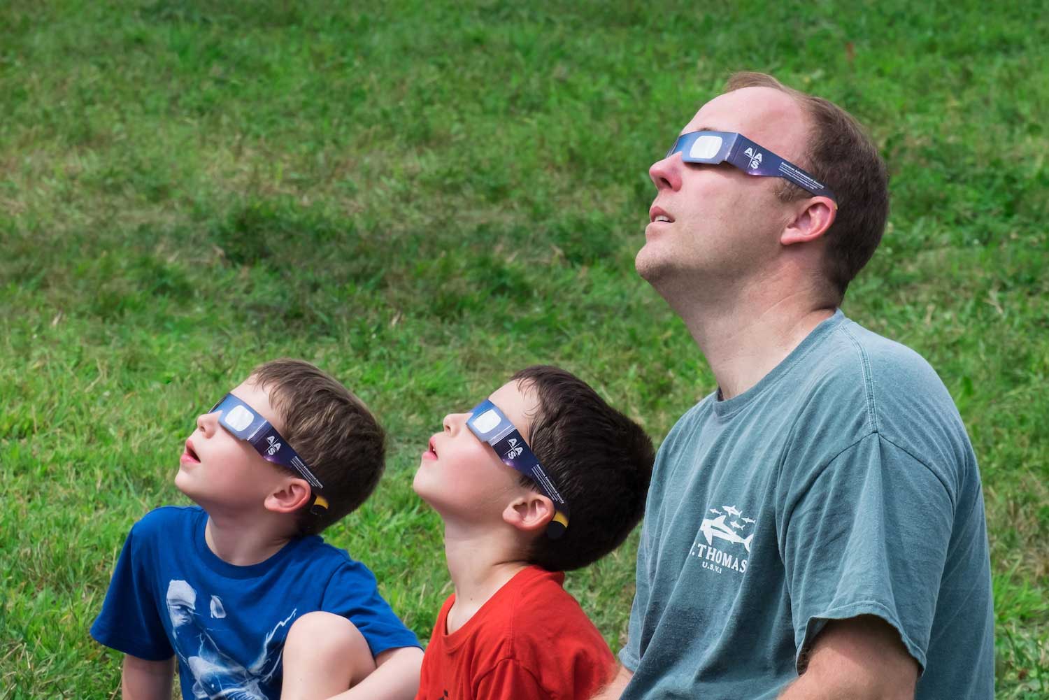 An adult and two children wearing solar viewing glasses while looking up at a solar eclipse.