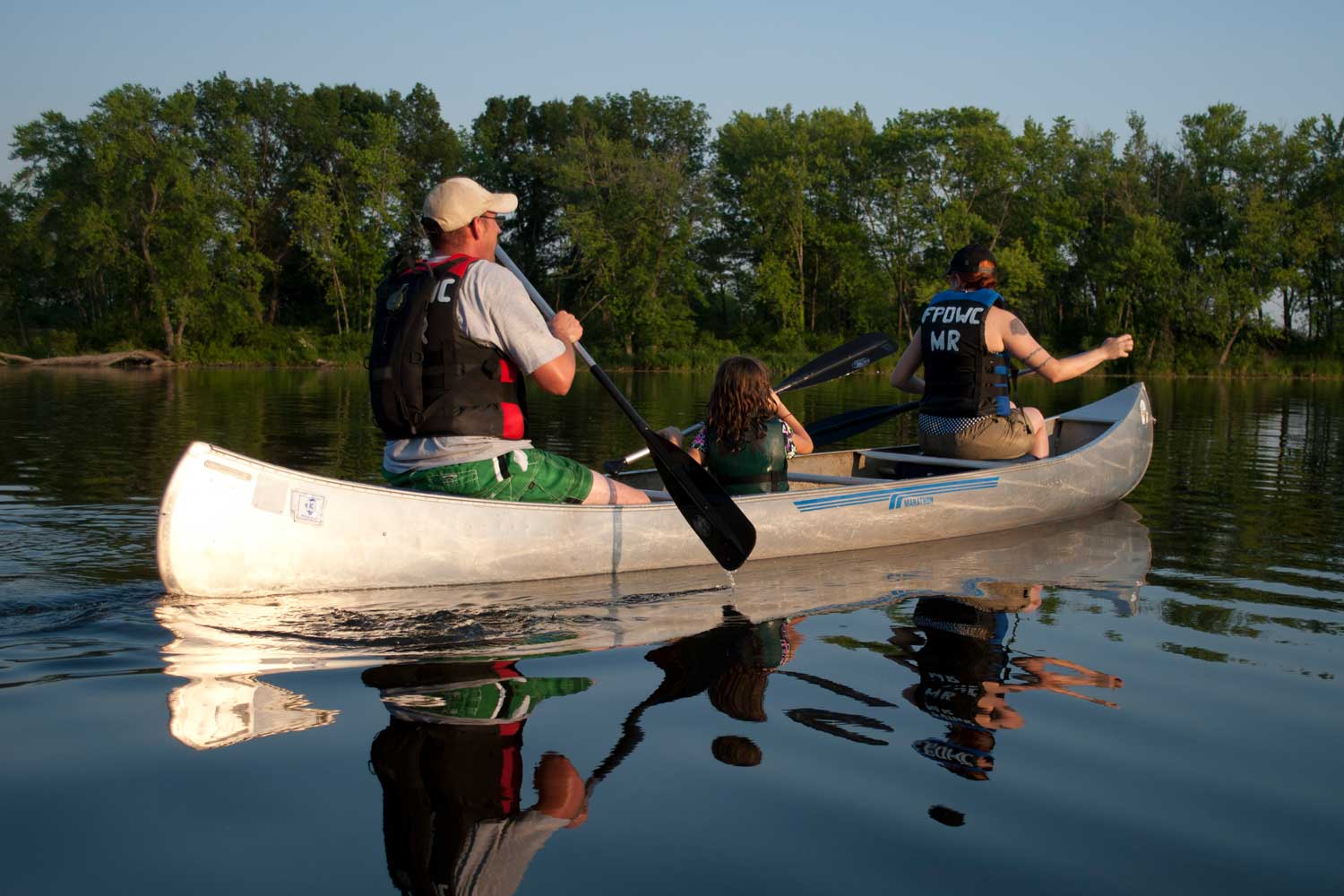 Three people in a canoe with the water and tree line in the background.