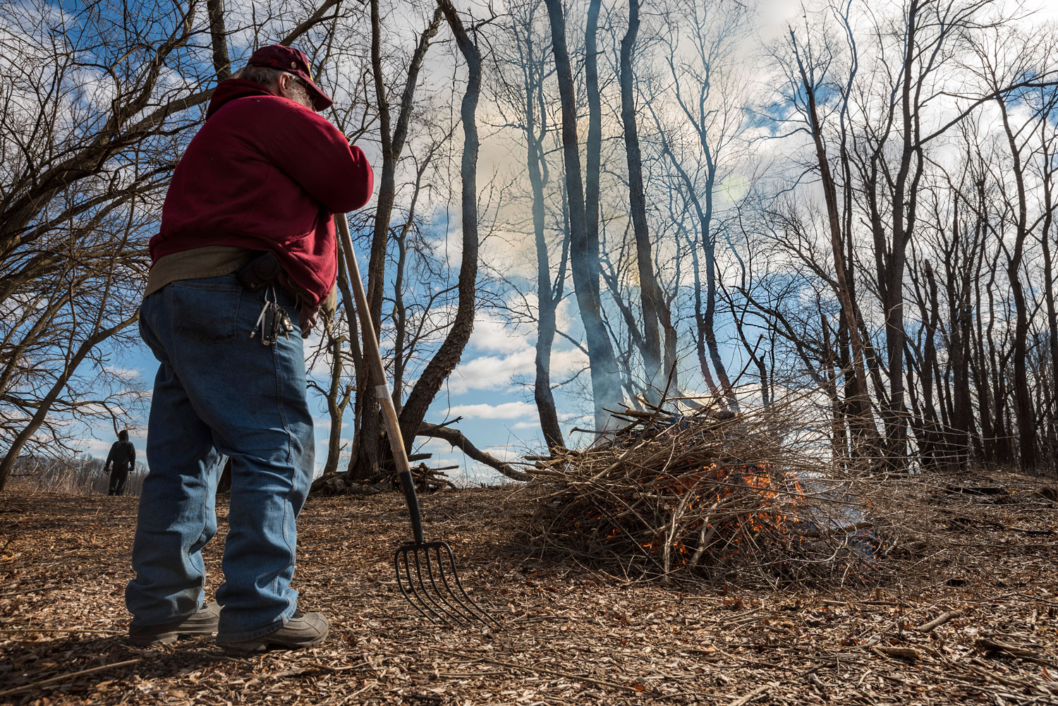 A person standing with a rake while watching a brush fire.