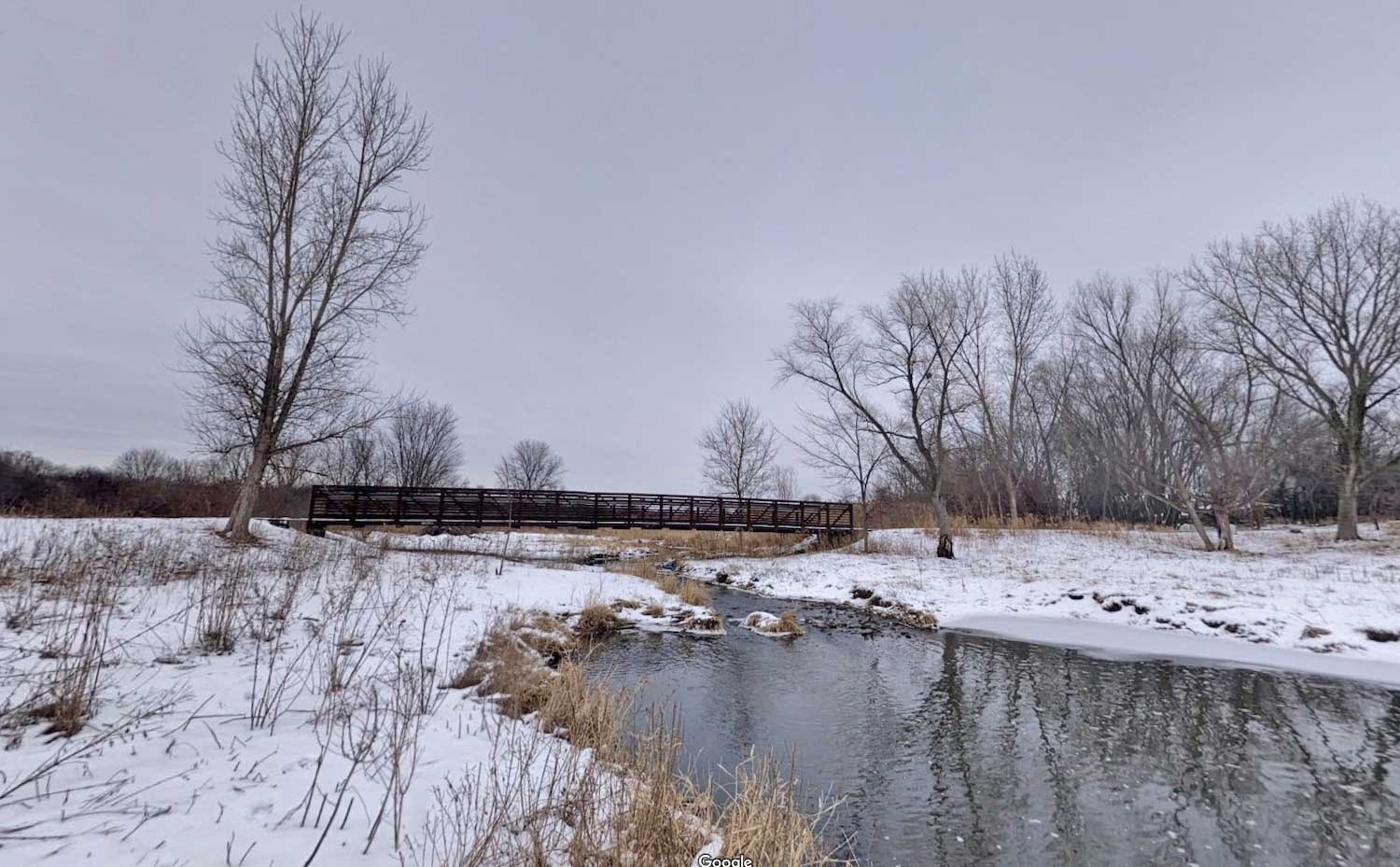 A creek surrounded by a light dusting of snow with a bridge in the background.