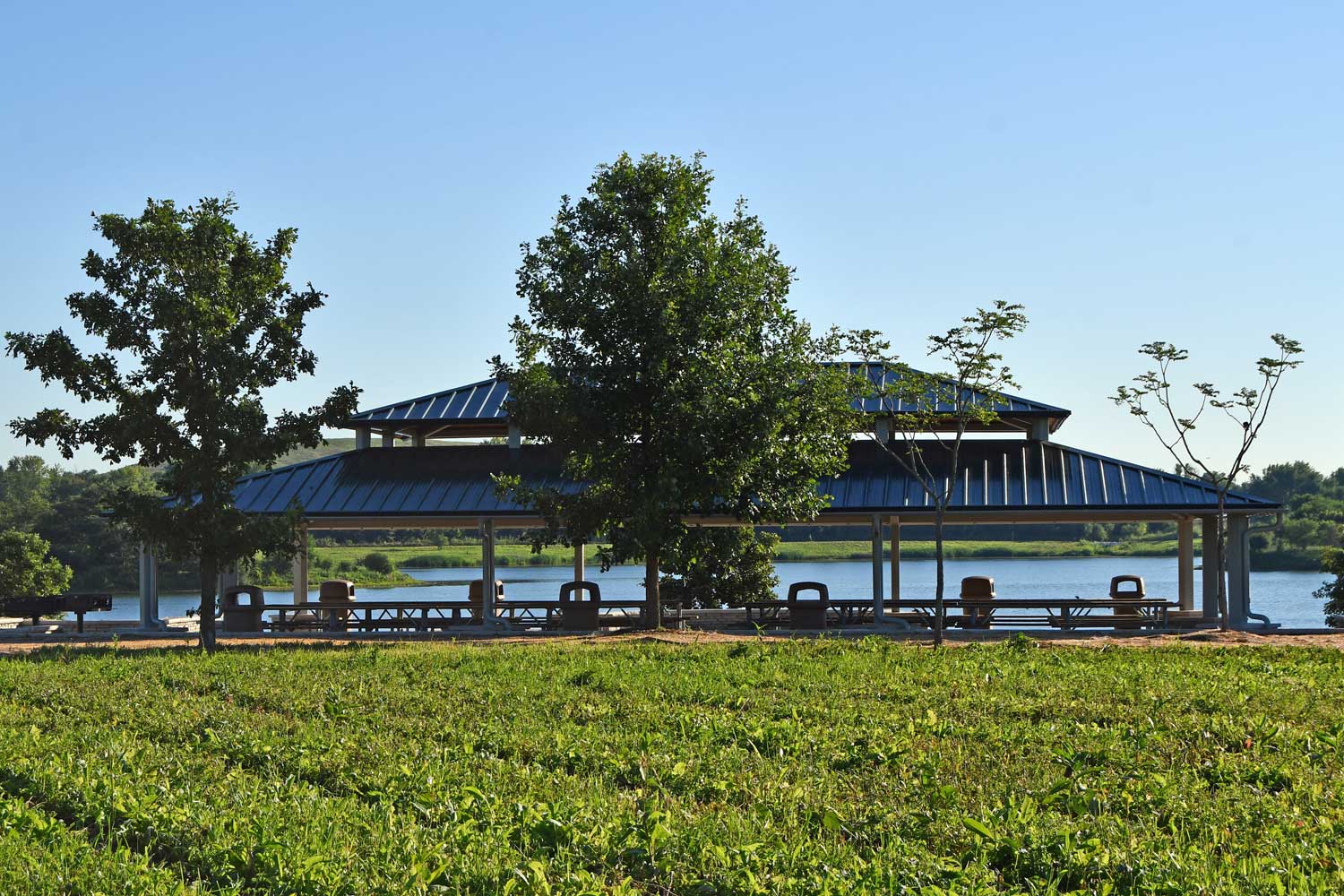 A grassy area with a large picnic shelter behind it and a lake providing a backdrop to the shelter.
