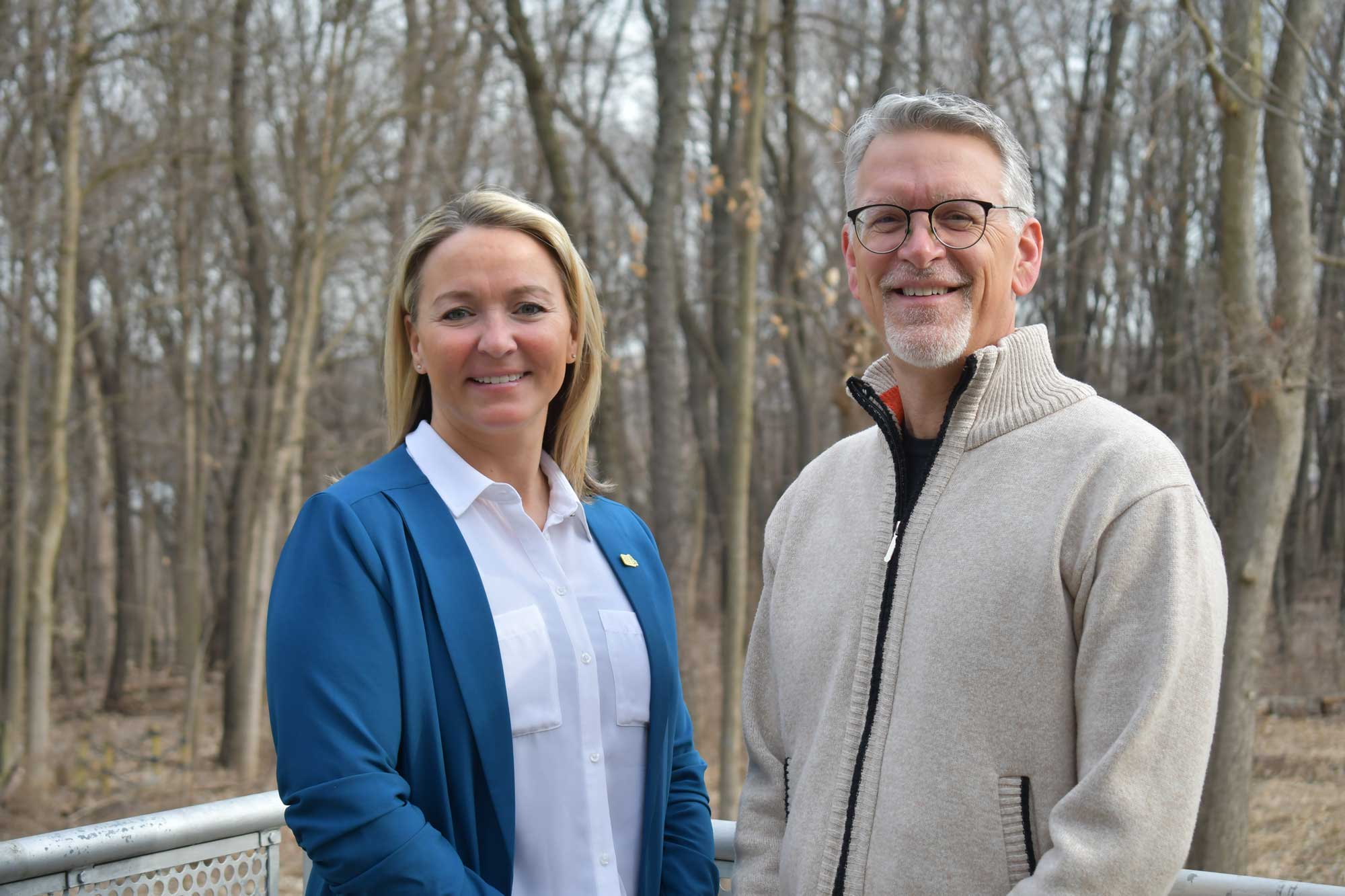 A man and a woman pose for a photo outside a nature center with woods in the background.