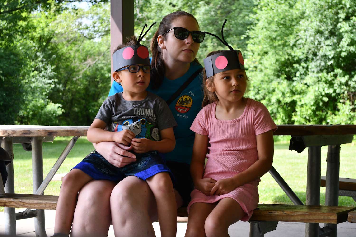 An adult and two kids sitting on the bench of a picnic table under a picnic shelter.