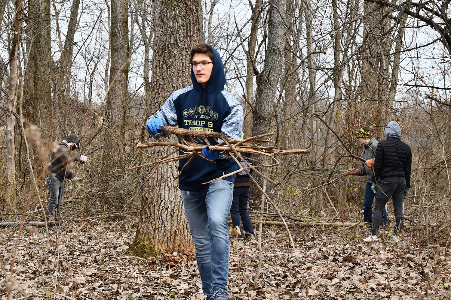 A person holding an armful of cut branches while three other people work on cutting brush in the background.