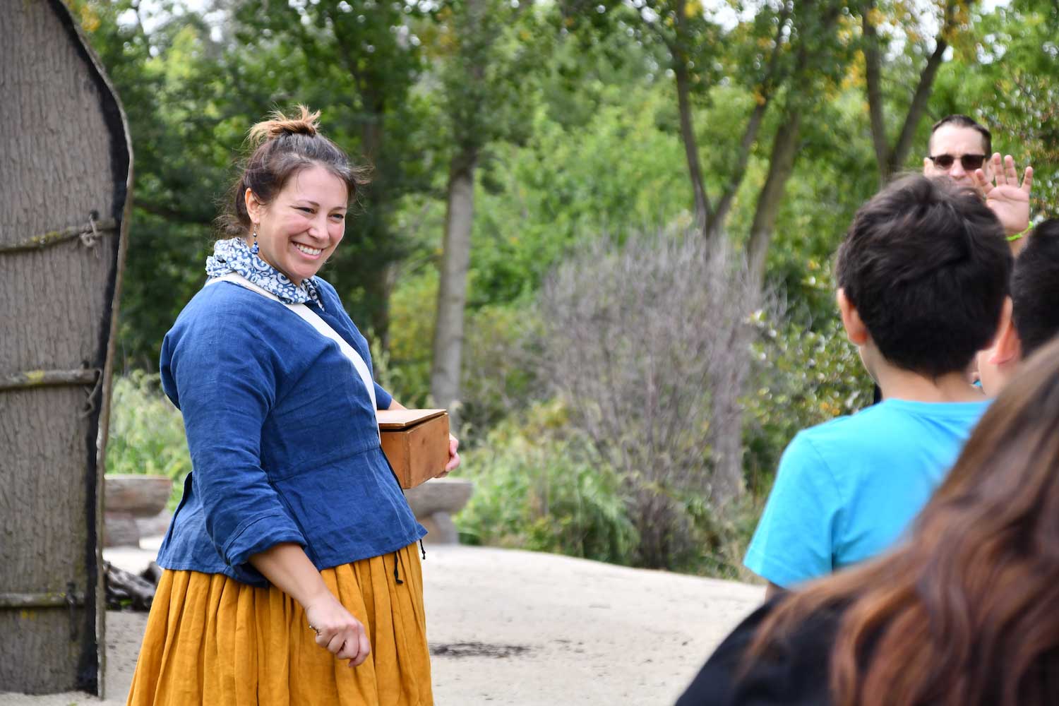 A person wearing traditional 18th-century clothing talking to a group of students while standing outside a replica Native American longhouse