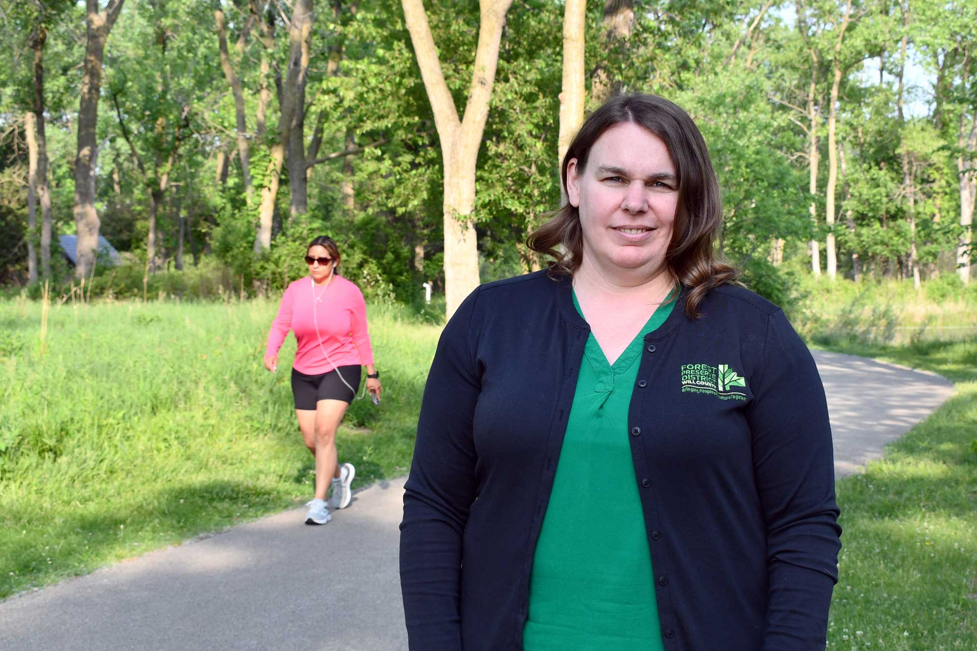 A woman poses for a photo on a trail while a runner passes by.