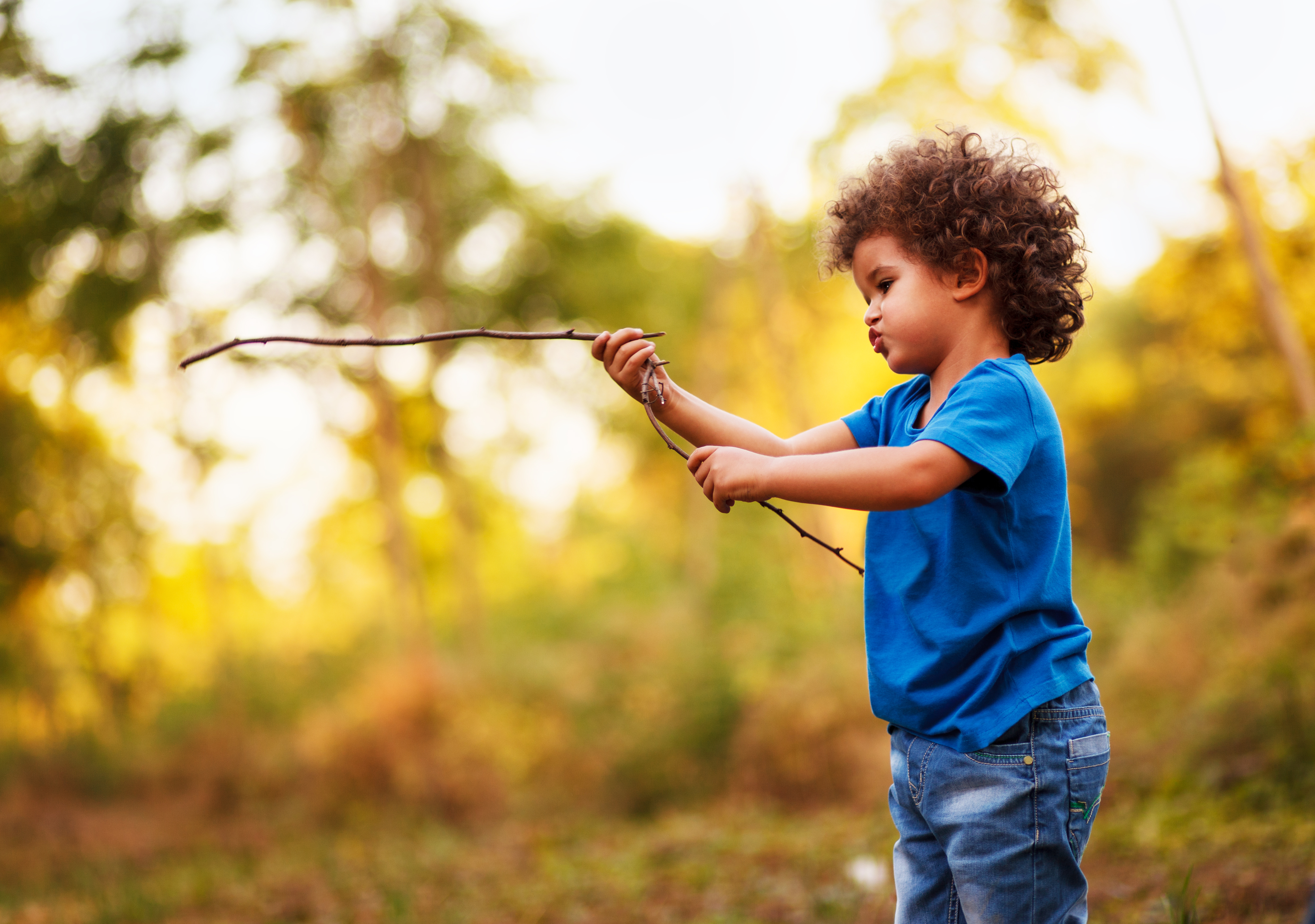 A young child holding two long sticks while standing on the ground with trees in the background.