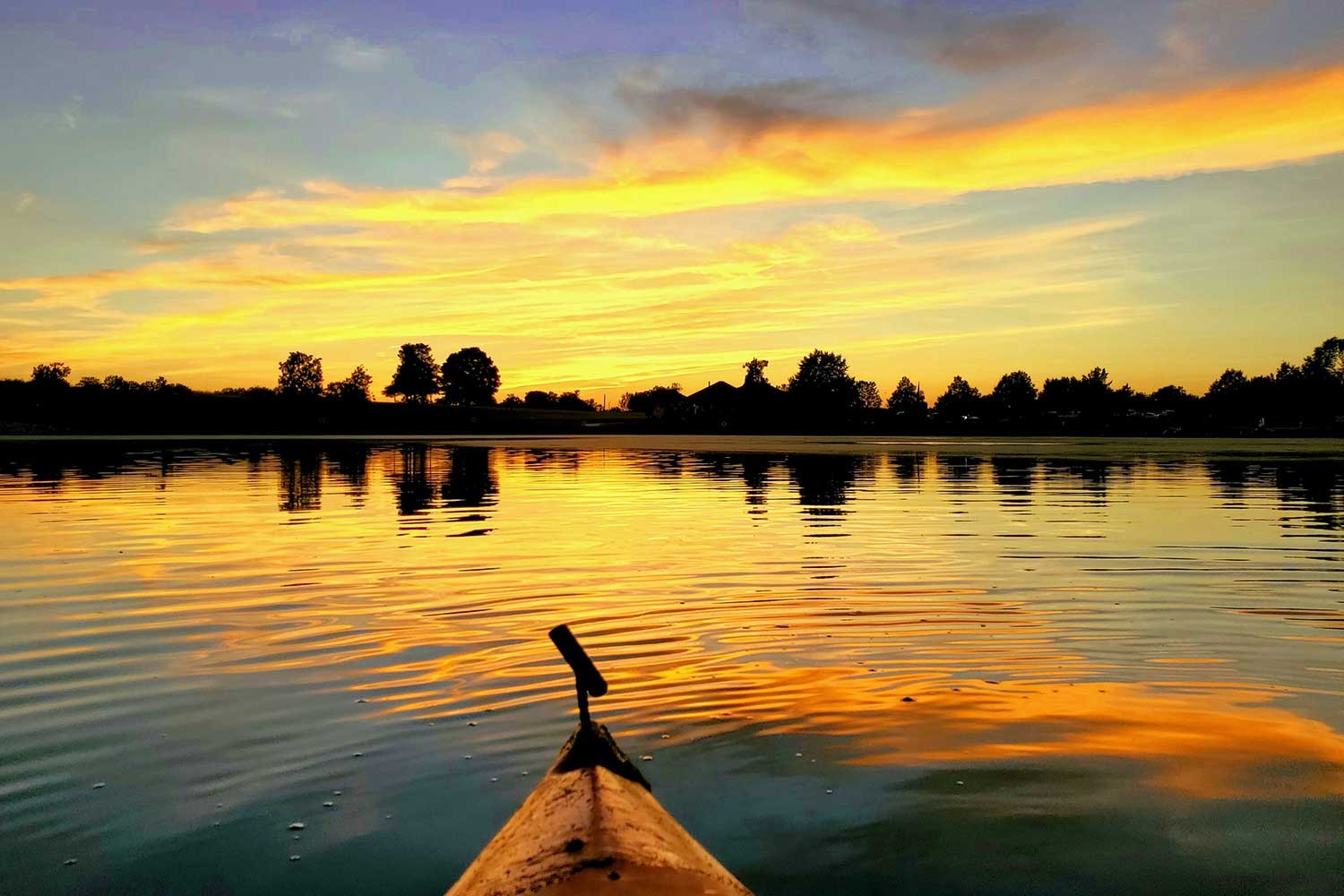 The tip of a kayak moving through the water with a colorful sunset of yellows and oranges.