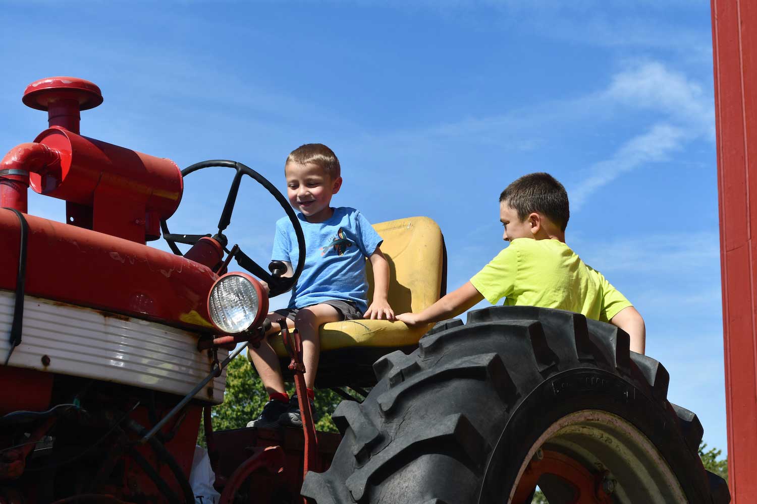 Two children on an antique tractor, with one sitting on the seat with a hand on the steering wheel and one standing to the side.