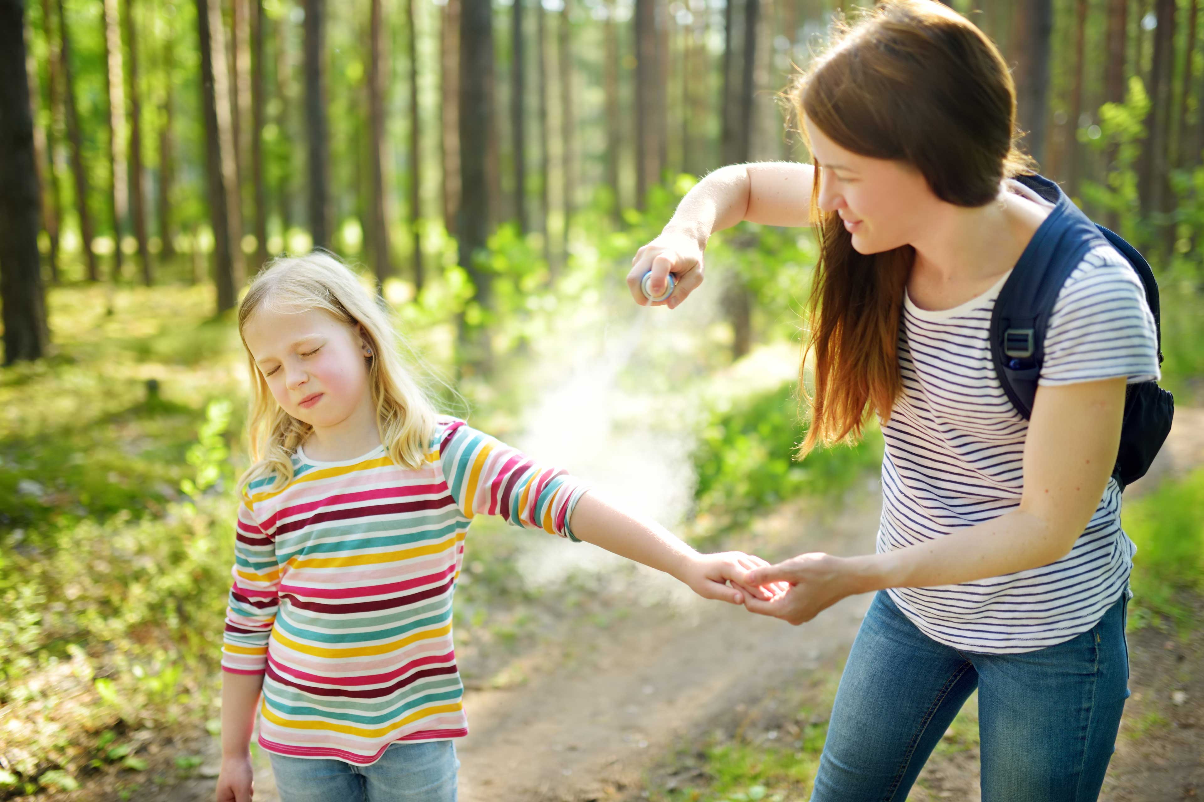 A woman spraying bug spray on a girl in the woods.