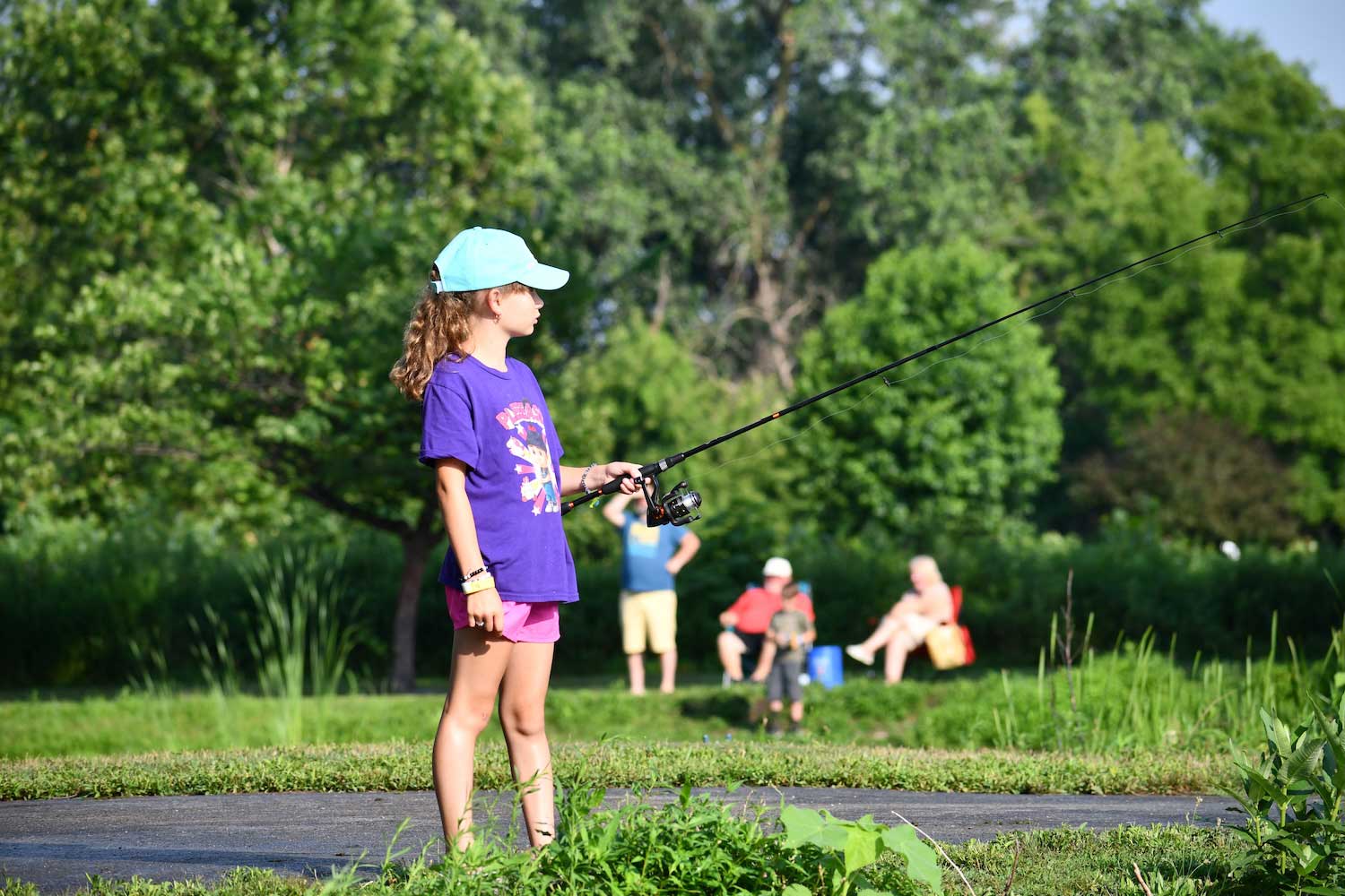 A child holding a fishing pole cast into the water while standing along a trail near a shoreline. People can be seen fishing in the background.