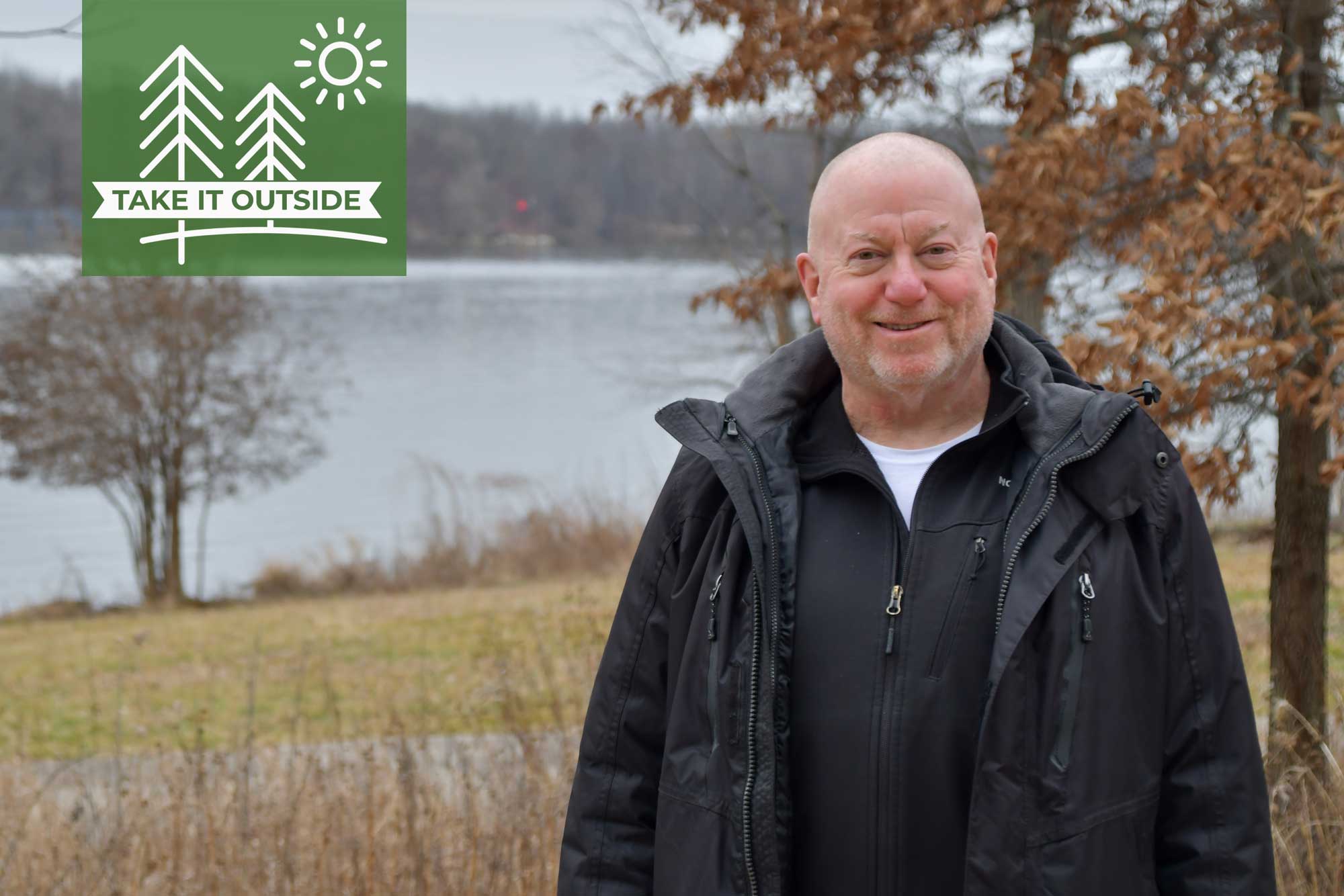 Man in black coat stands along trail with river in the background.