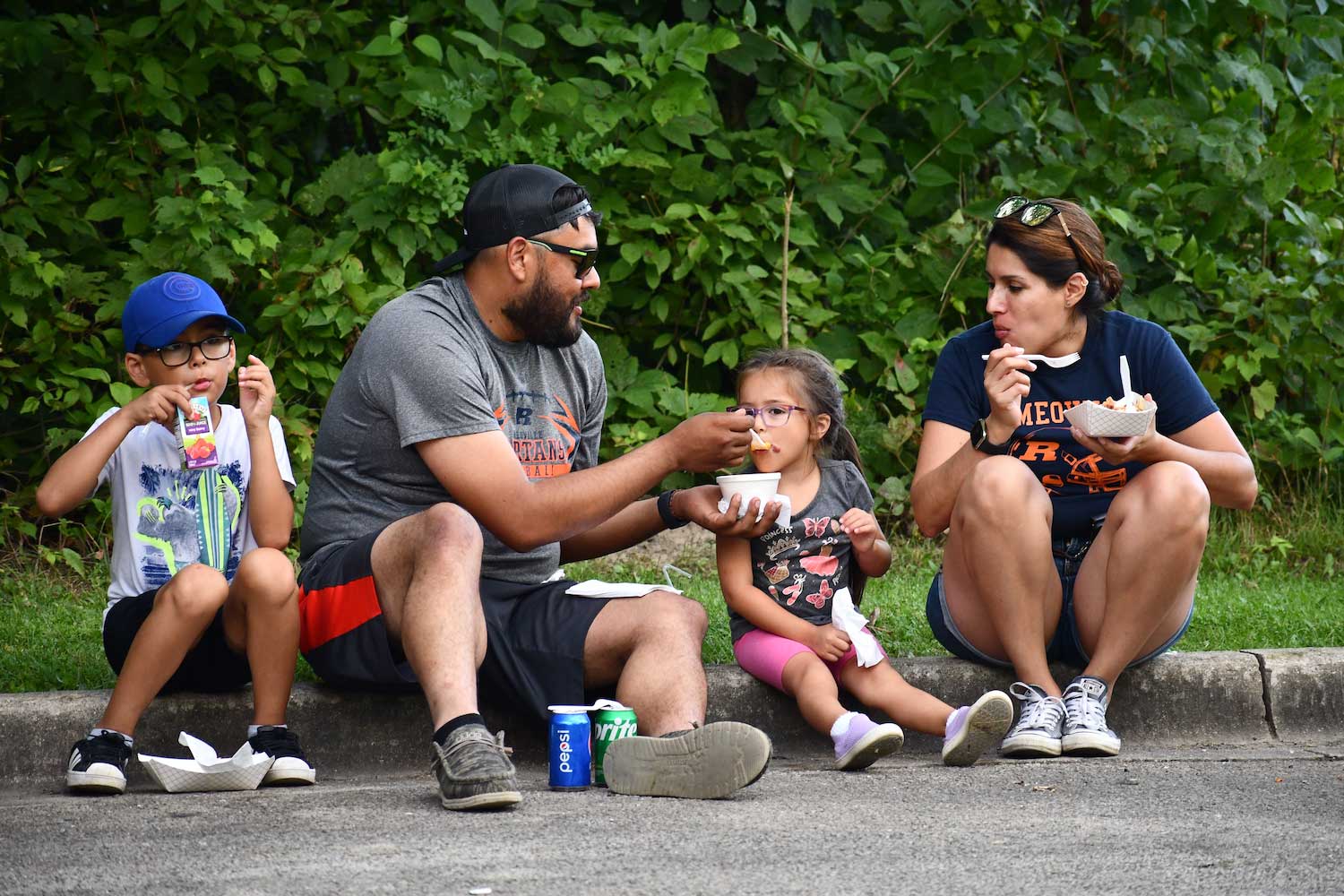 A family of four sitting on a curb while enjoying food truck fare.