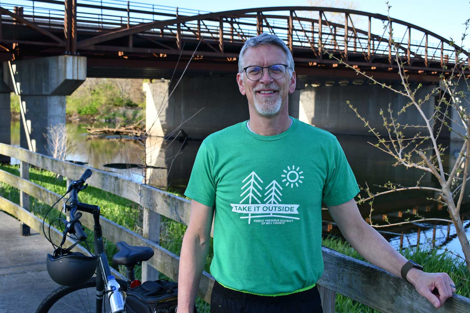 A man next to a bike standing along a trail that runs along a river with a bridge in the background.