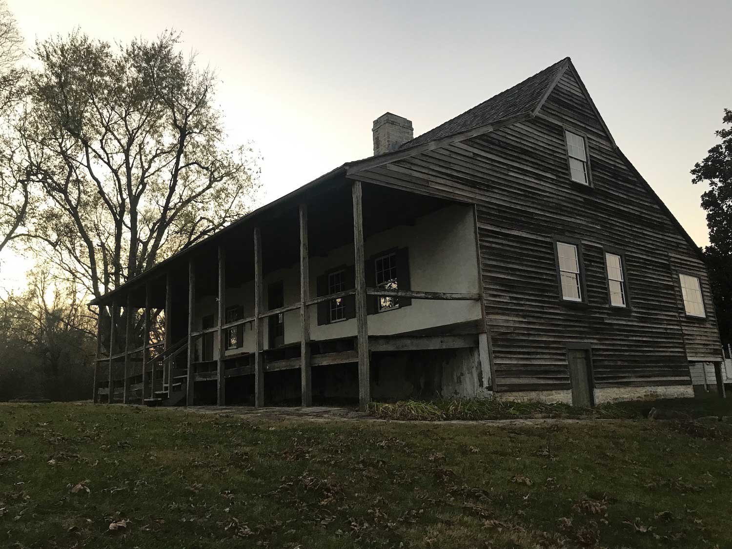 An old house with a wooden porch with bare trees and the setting sun in the background.
