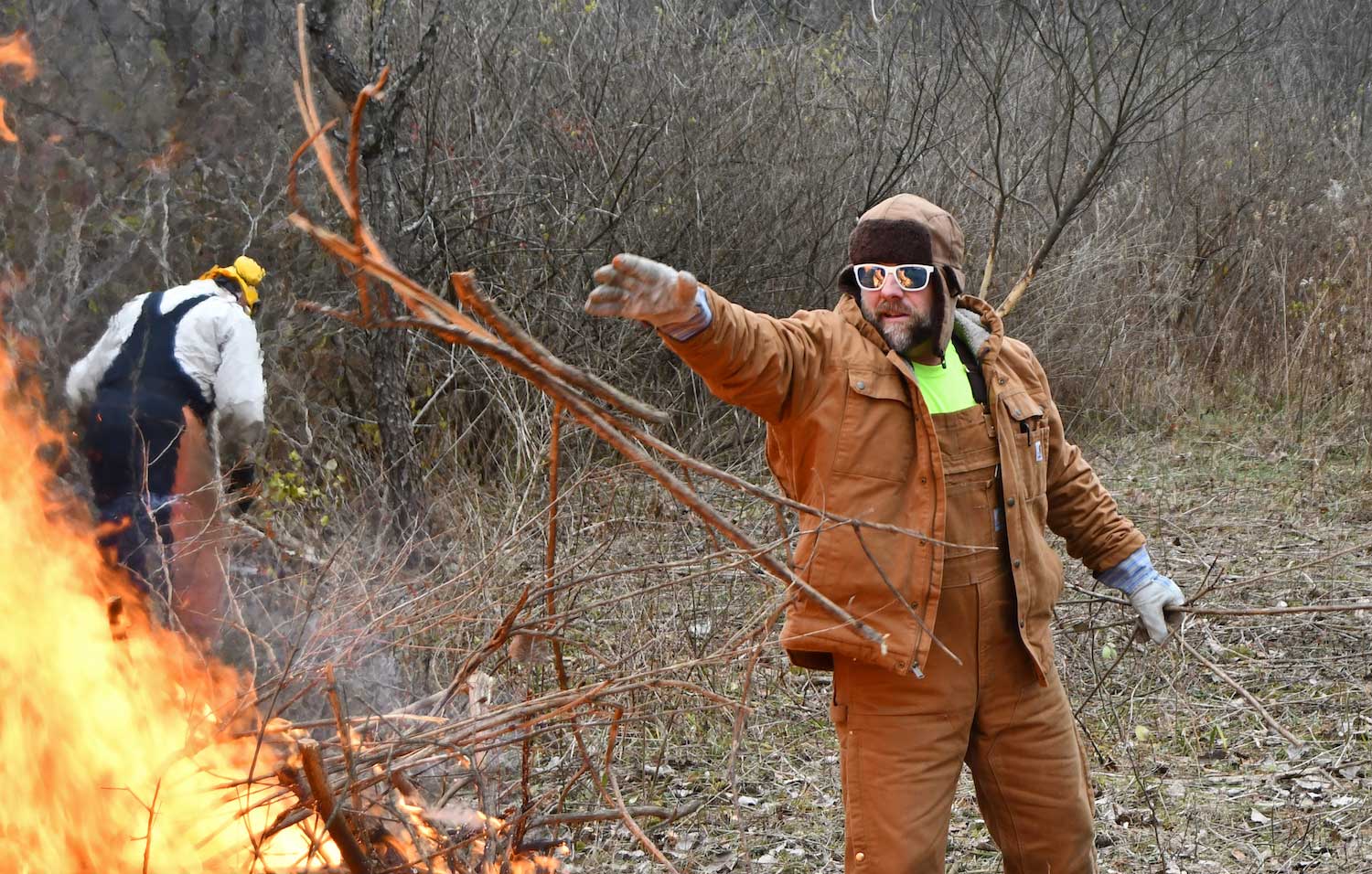 A person throwing a large branch on a burning brush pile.