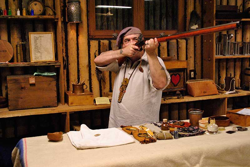 A person holding a replica weapon while standing behind a table in a replica traders' cabin.