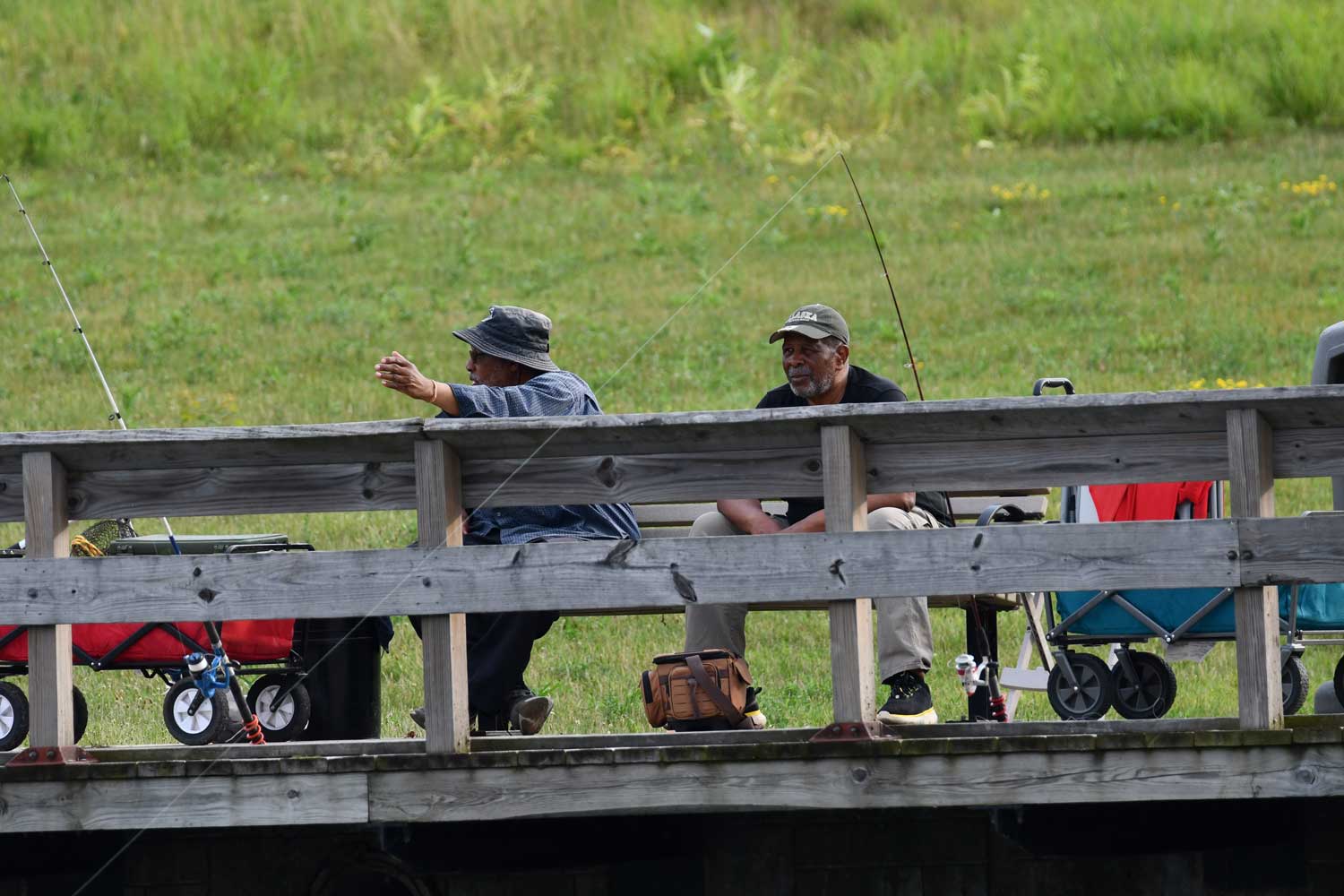 Two people sitting on a bench behind a wooden railing. Cast fishing poles are leaned up against the railing.