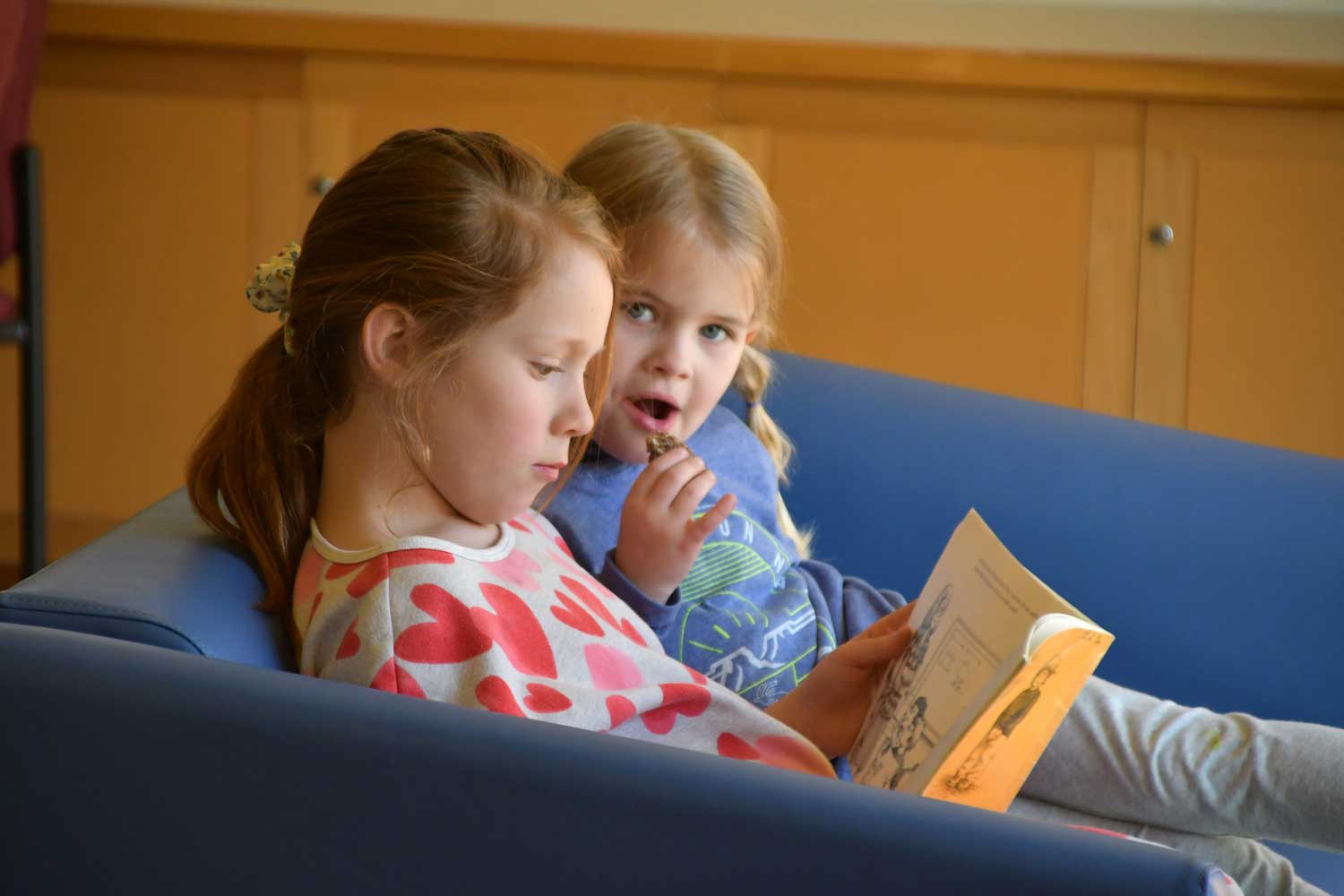 Two children sitting in a couch looking at a book.