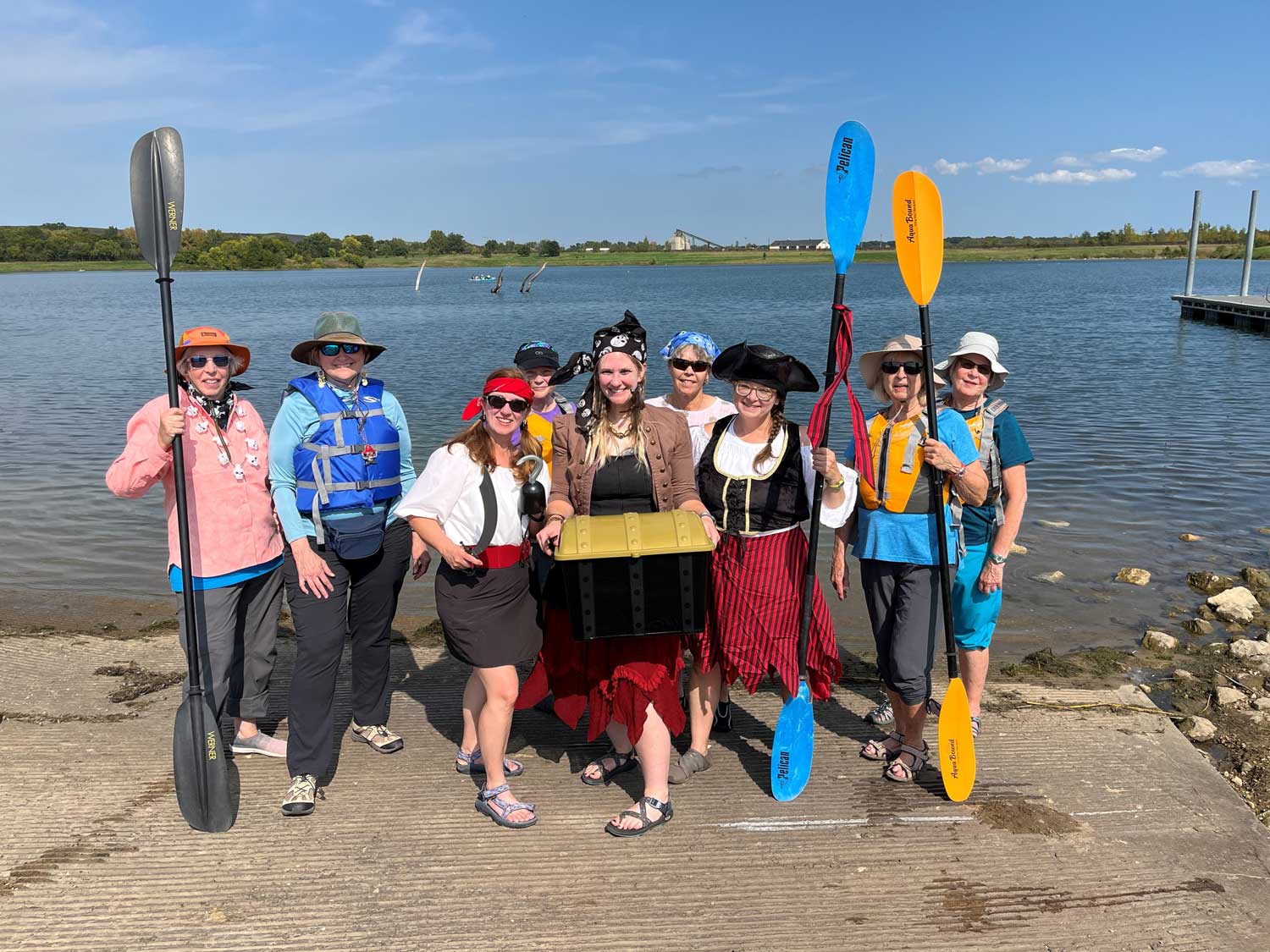 A group of people dressed in pirate garb and holding kayak paddles standing in front of a lake.