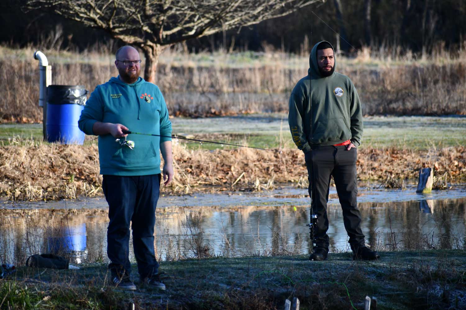 Two people standing along a shoreline with a pond in the background. One person is holding a fishing pole.