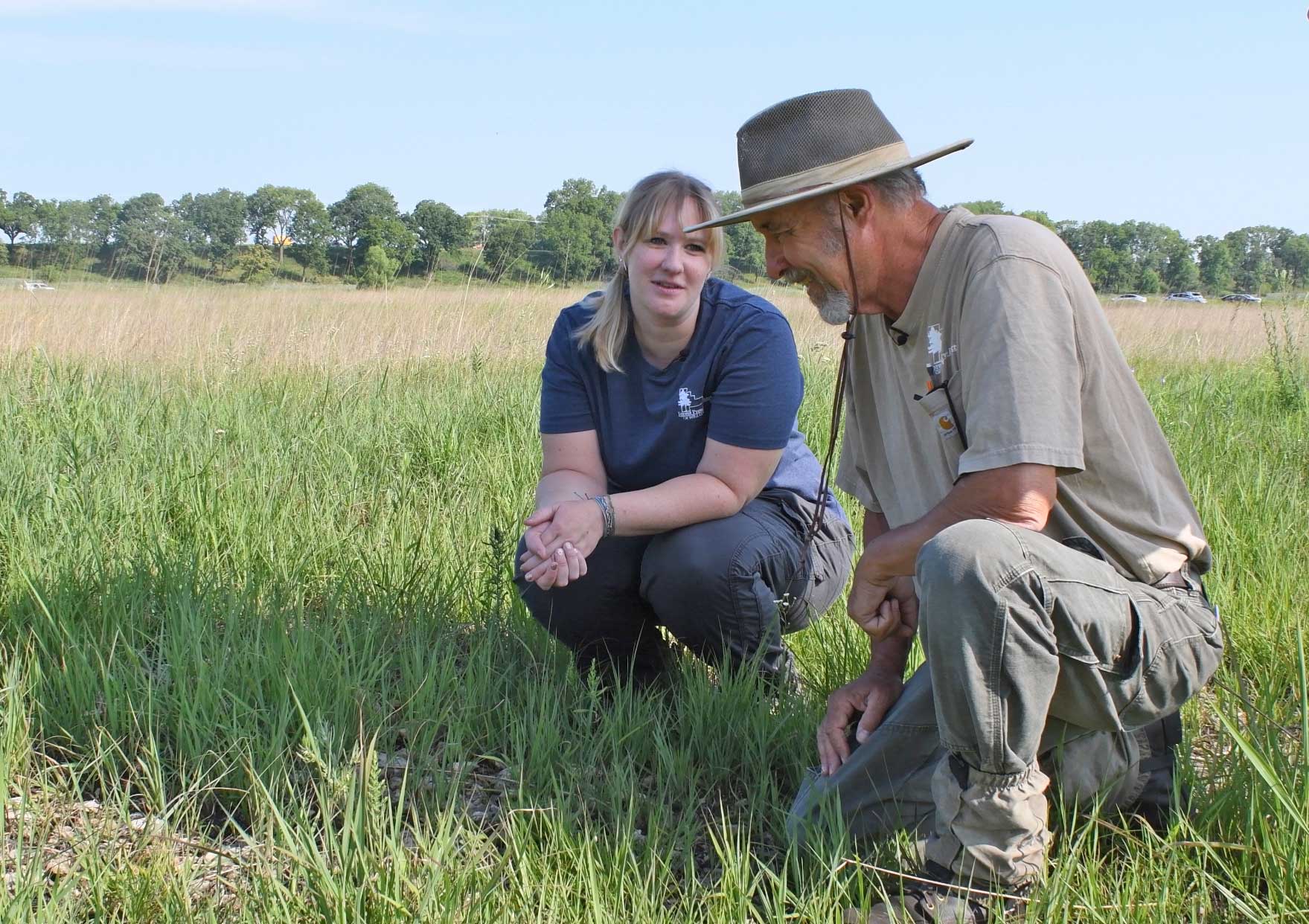 A man and woman in a kneel down in a prairie to look at a plant.