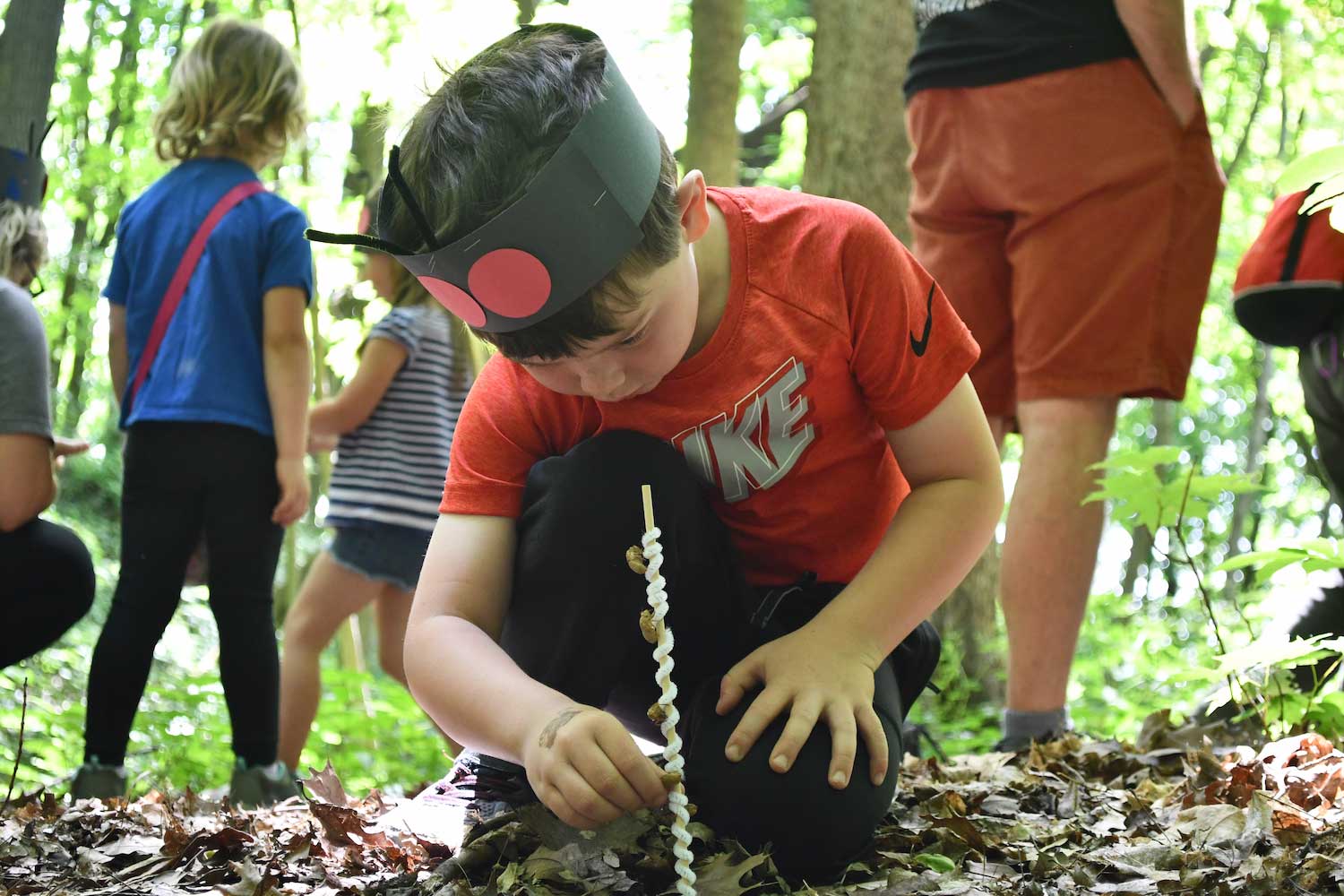 A young child kneeling on the ground while holding up a stick in the dirt. More children are in the background.