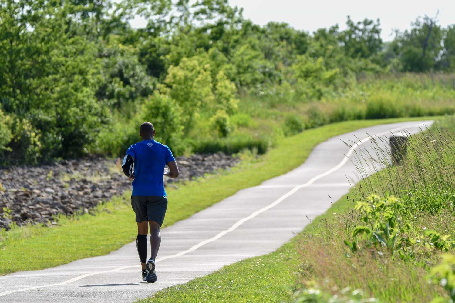 A man running along a paved trail lined with trees and grasses.