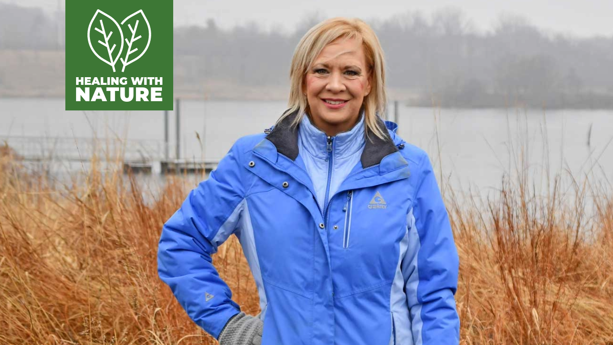 A woman in a blue jacket stands in a forest preserve with water in the background.