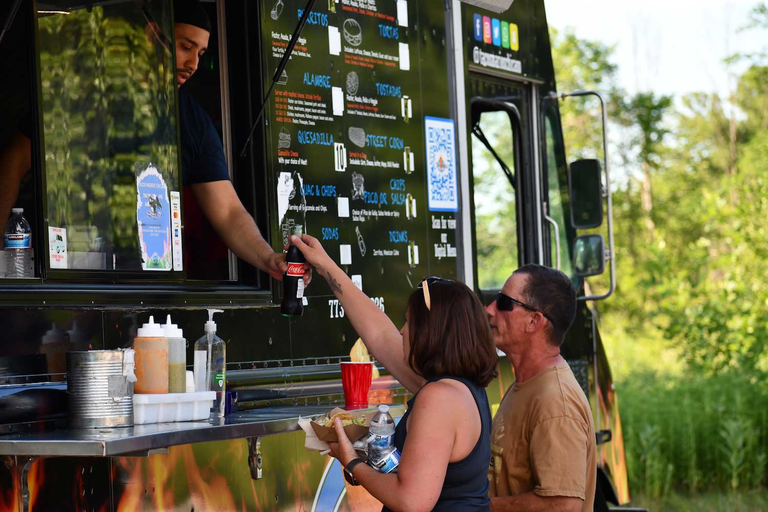 Two people standing at the window of a food truck while an employee passes them a drink.
