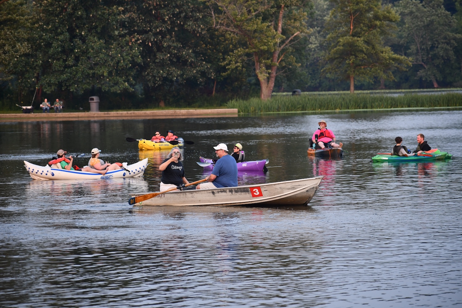 People in kayaks, canoes and rowboats on a lake on a summer evening.