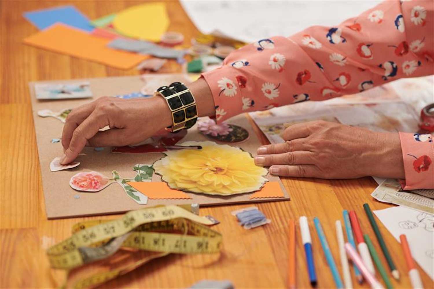 A person making a collage at a wooden table. The table is covered with art supplies.