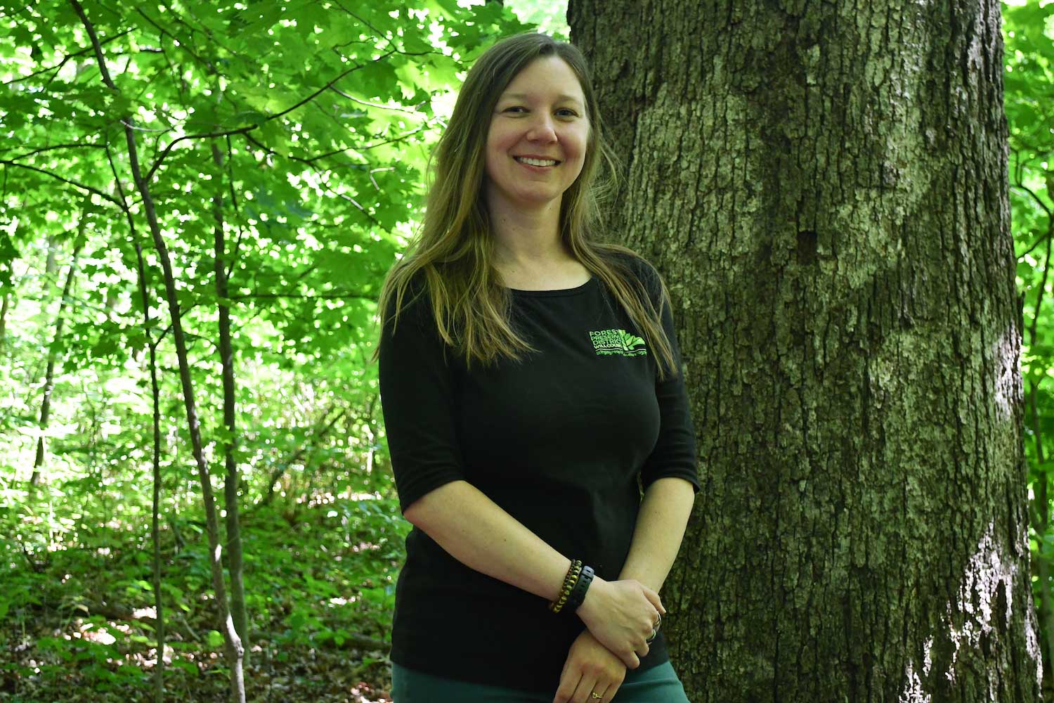 A person standing next to a large tree trunk in a forest.