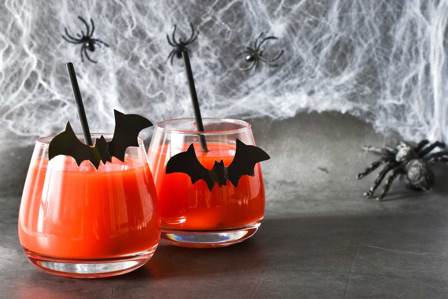 Two bright orange cocktails/mocktails on a table. The glasses are decorated with bats and there are fake spider webs in the background.