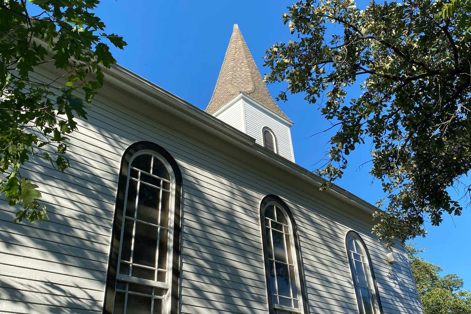 A closeup view on an old church looking up at its steeple with trees in the foreground and blue sky in the background.